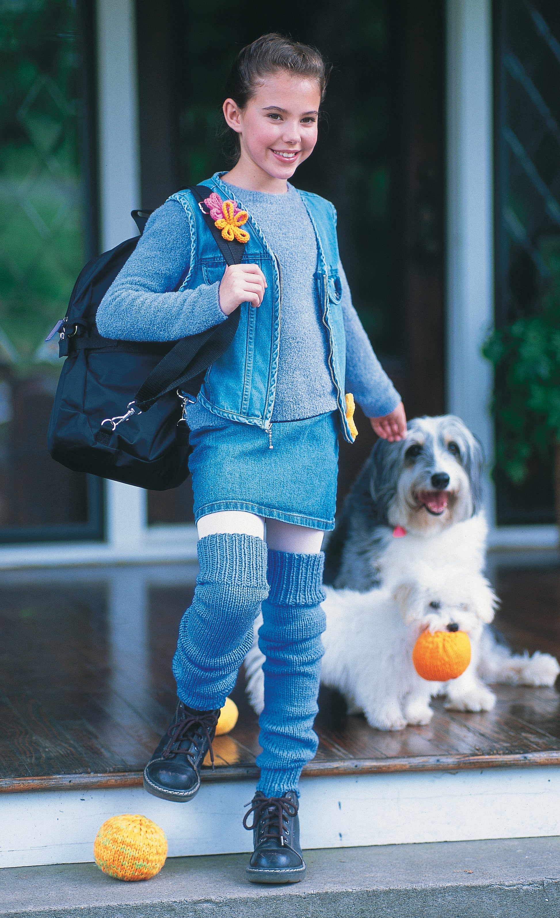 A smiling child in denim and blue leg warmers—ideal for Leisure Arts’ Cool Stuff for Kids Teach Me to Knit—12 Projects—stands on a porch with a backpack, two fluffy dogs, and small orange pumpkins on the steps.