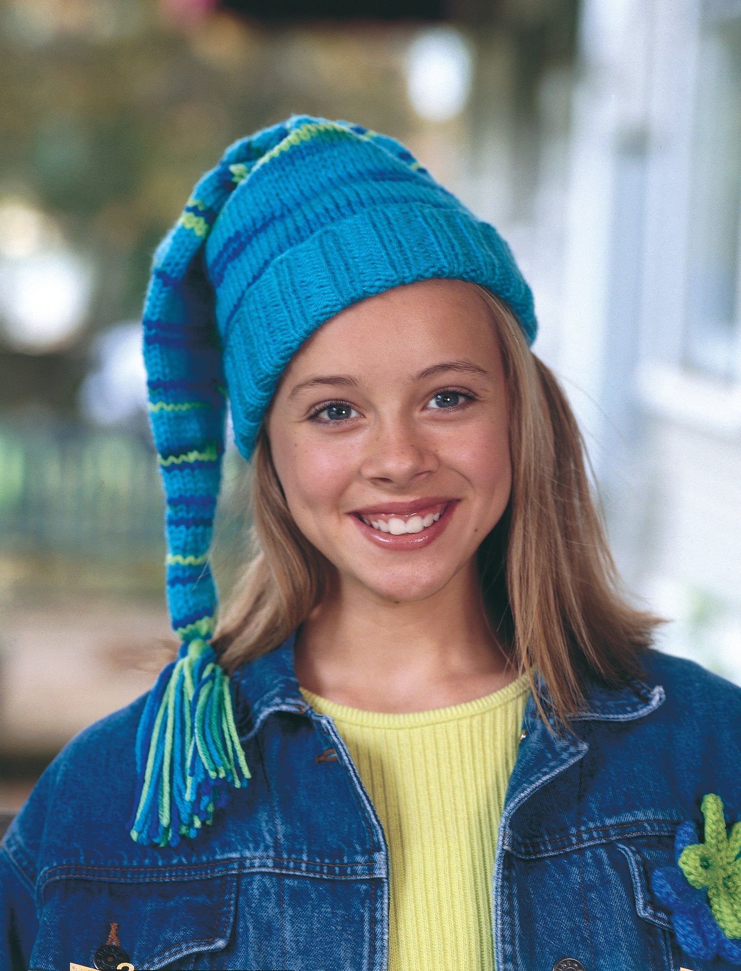 A smiling young girl with straight blonde hair wears a long blue and green knit hat, made from Leisure Arts' "Cool Stuff for Kids Teach Me to Knit—12 Projects," paired with a denim jacket and yellow shirt outdoors in soft natural light.