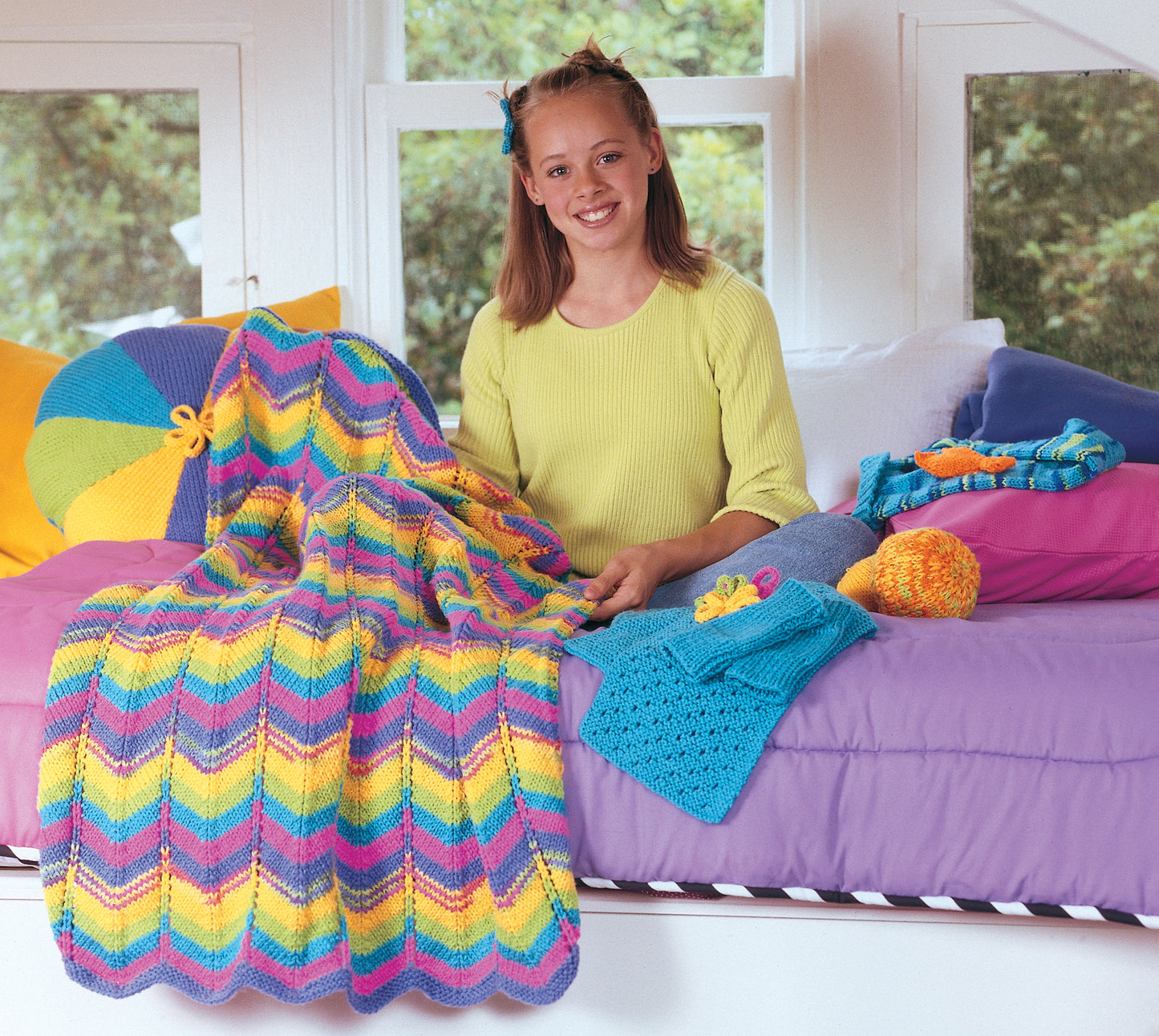 A smiling girl in a yellow top sits on a bed with colorful chevron bedding, surrounded by yarn and projects from the Leisure Arts Cool Stuff for Kids Teach Me to Knit—12 Projects kit. Knitted items, a striped pillow, and greenery outside complete the scene.