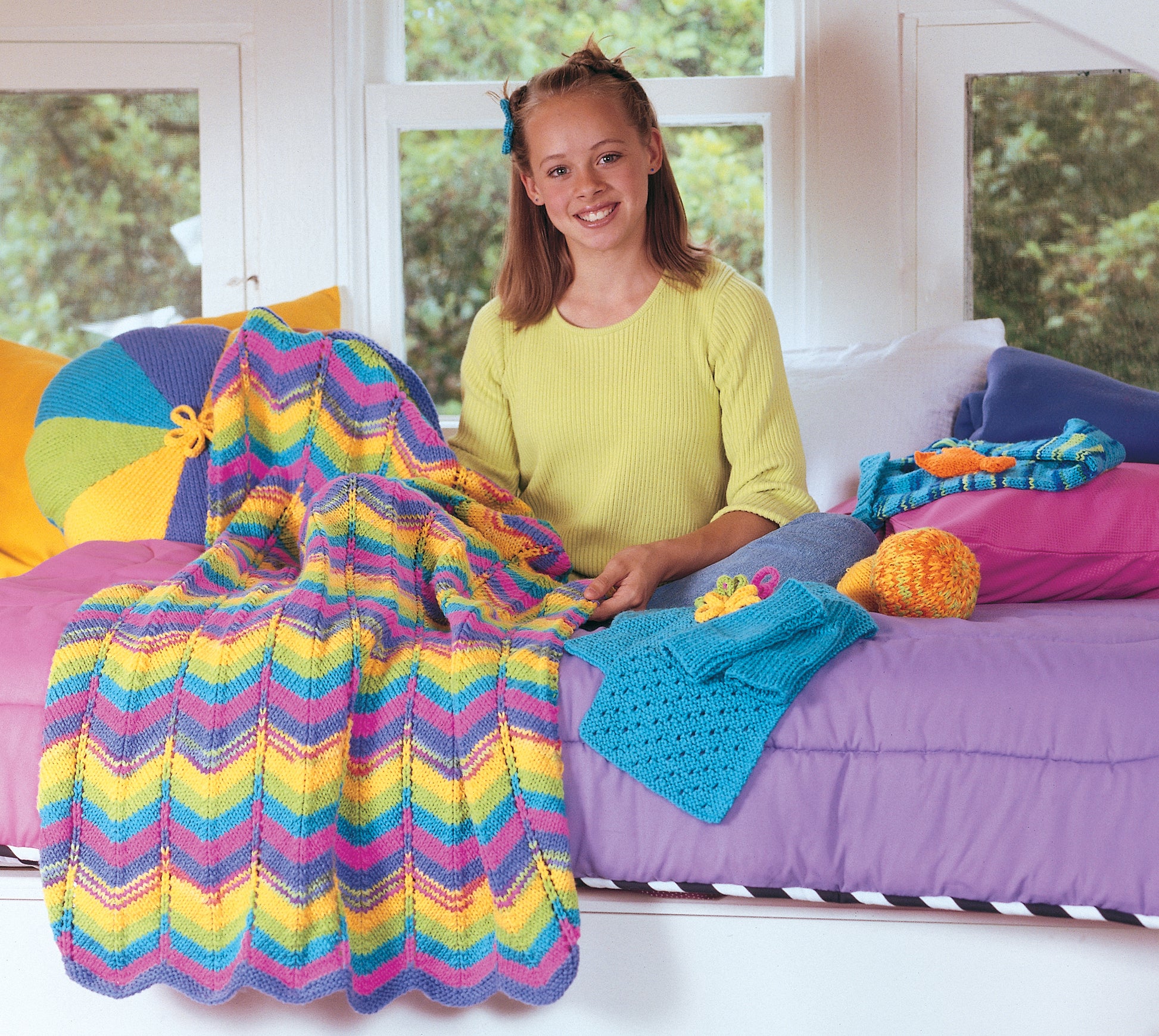 A smiling girl in a yellow top sits on a bed with colorful chevron bedding, surrounded by yarn and projects from the Leisure Arts Cool Stuff for Kids Teach Me to Knit—12 Projects kit. Knitted items, a striped pillow, and greenery outside complete the scene.