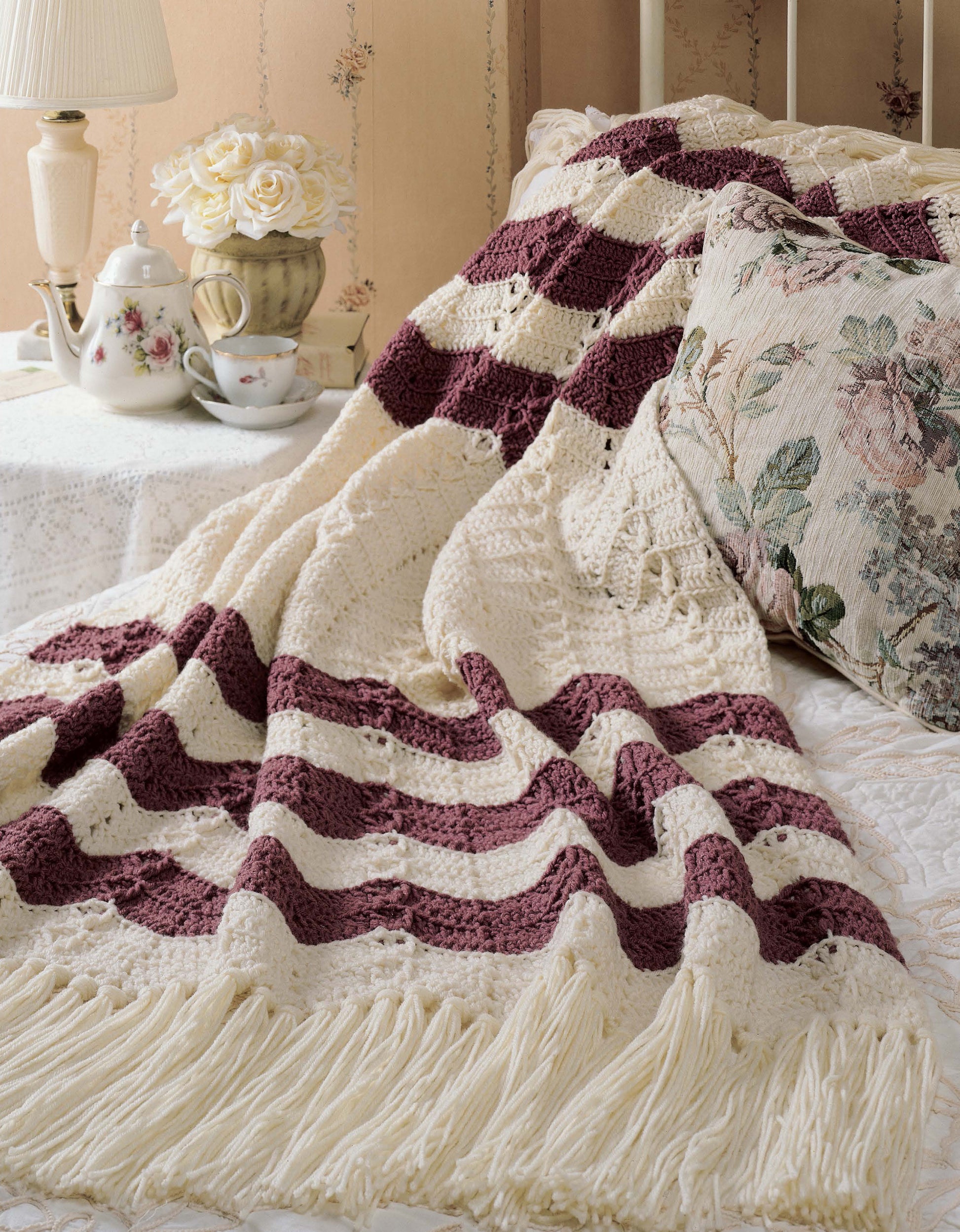 A cream and burgundy ripple afghan from Leisure Arts' "40 Favorite Ripple Afghans" adds charm to the bed, paired with a floral pillow. Nearby, a teapot, cup, and white roses sit atop a lace-draped bedside table.