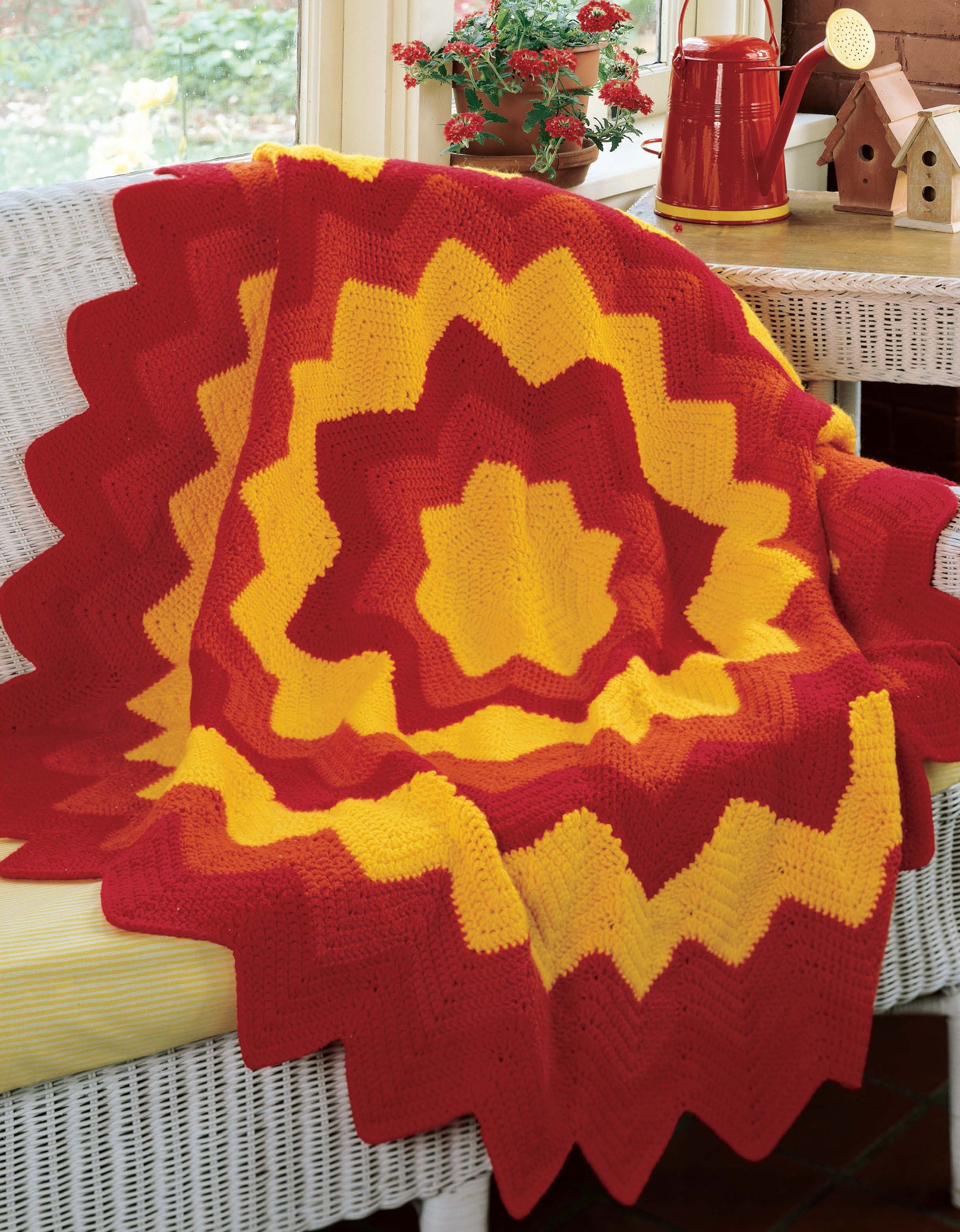 A red and yellow star-patterned crocheted blanket from Leisure Arts' "40 Favorite Ripple Afghans" is displayed on a white wicker bench by a window, with potted plants, birdhouses, and a red teapot in the background.