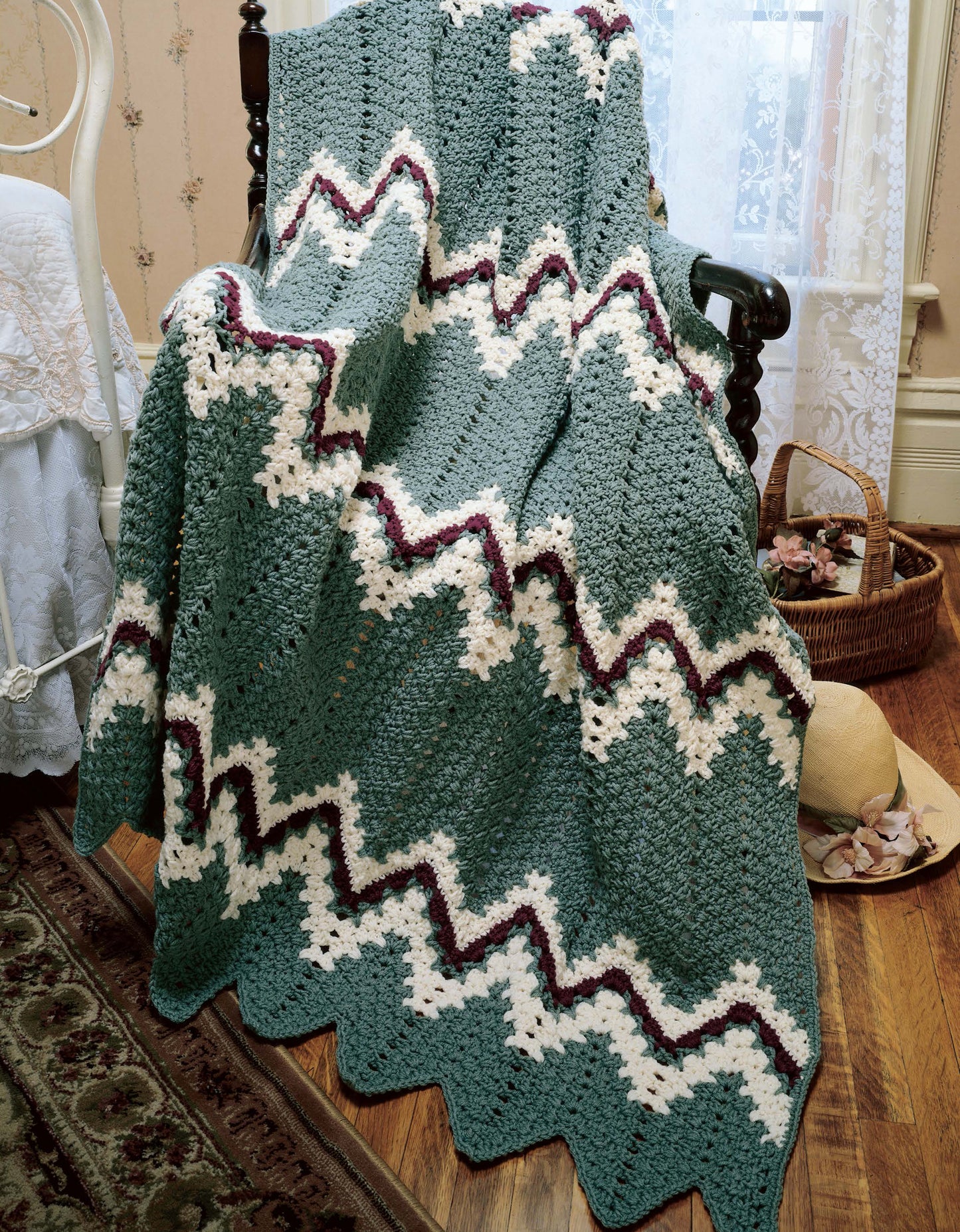 A green, white, and burgundy crocheted zigzag afghan from "40 Favorite Ripple Afghans" by Leisure Arts drapes over a wooden chair in a vintage room with lace curtains, a basket, hat, and wooden floors visible.