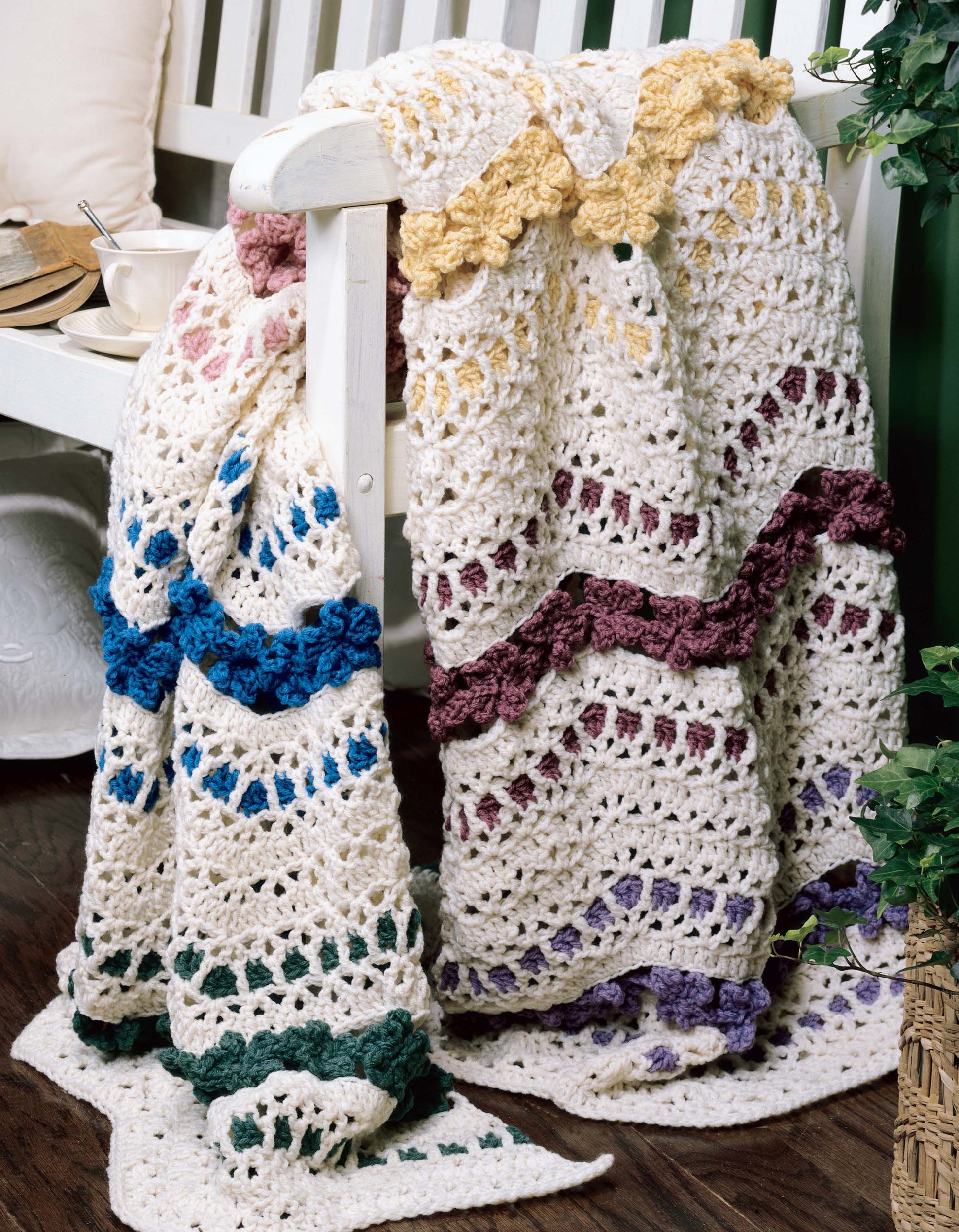 A vibrant ripple afghan from "40 Favorite Ripple Afghans" by Leisure Arts, in yellow, purple, blue, green, and pink, drapes over a white bench near a teacup, books, and a leafy plant. Featured in Crochet With Heart Magazine.