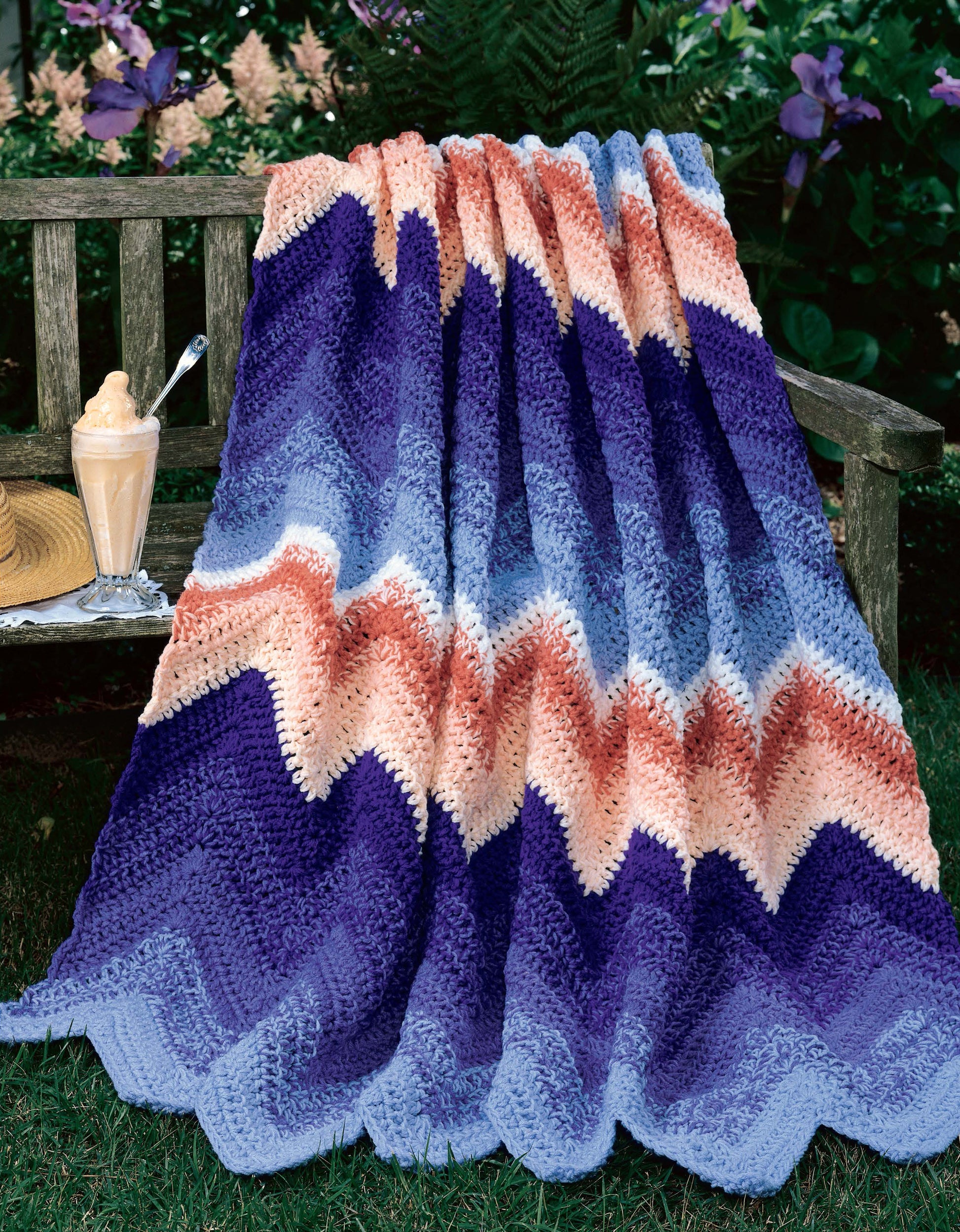 A crocheted blanket with zigzag stripes in blue, purple, peach, and beige from Leisure Arts’ 40 Favorite Ripple Afghans is draped over a wooden bench outdoors; purple flowers bloom behind a glass of creamy iced coffee.
