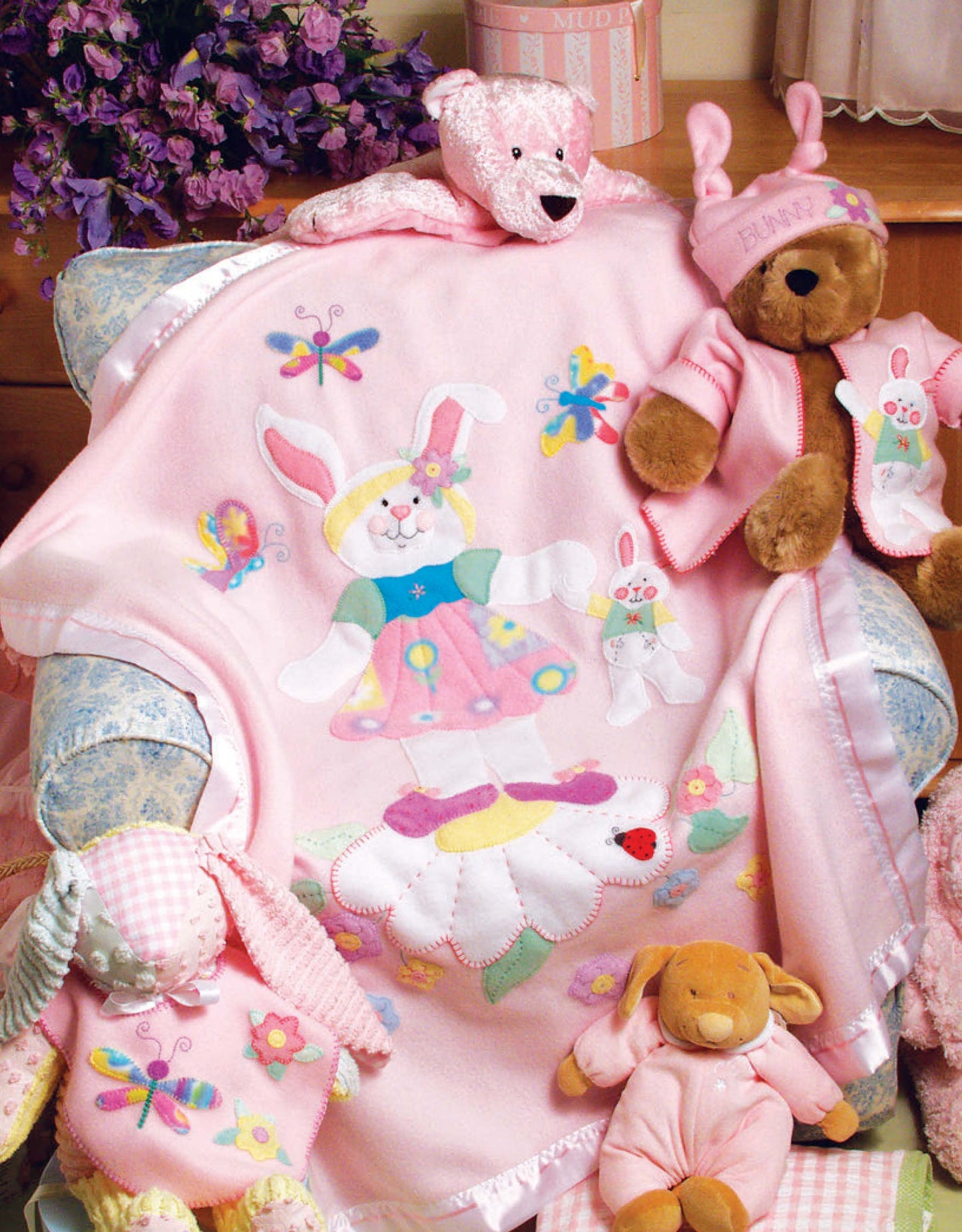 A comfy armchair displays a pink ballerina bunny blanket by Linda Gillum, with pink plush bears and a dog nearby, plus purple flowers. From "Baby Quilts and More—18 Projects" by Leisure Arts (Print ISBN 9781601407016).