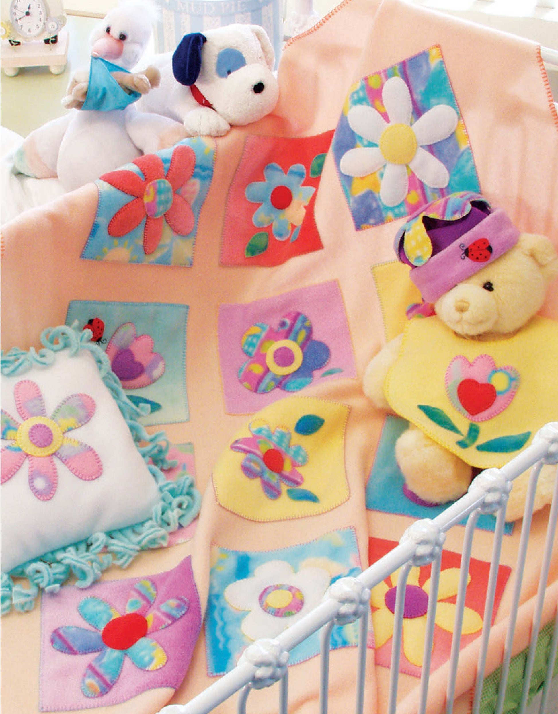 A pastel baby blanket with appliqué flower designs from Leisure Arts’ "Baby Quilts and More—18 Projects" is draped over a crib, surrounded by plush bears and a flower pillow. Print ISBN 9781601407016.