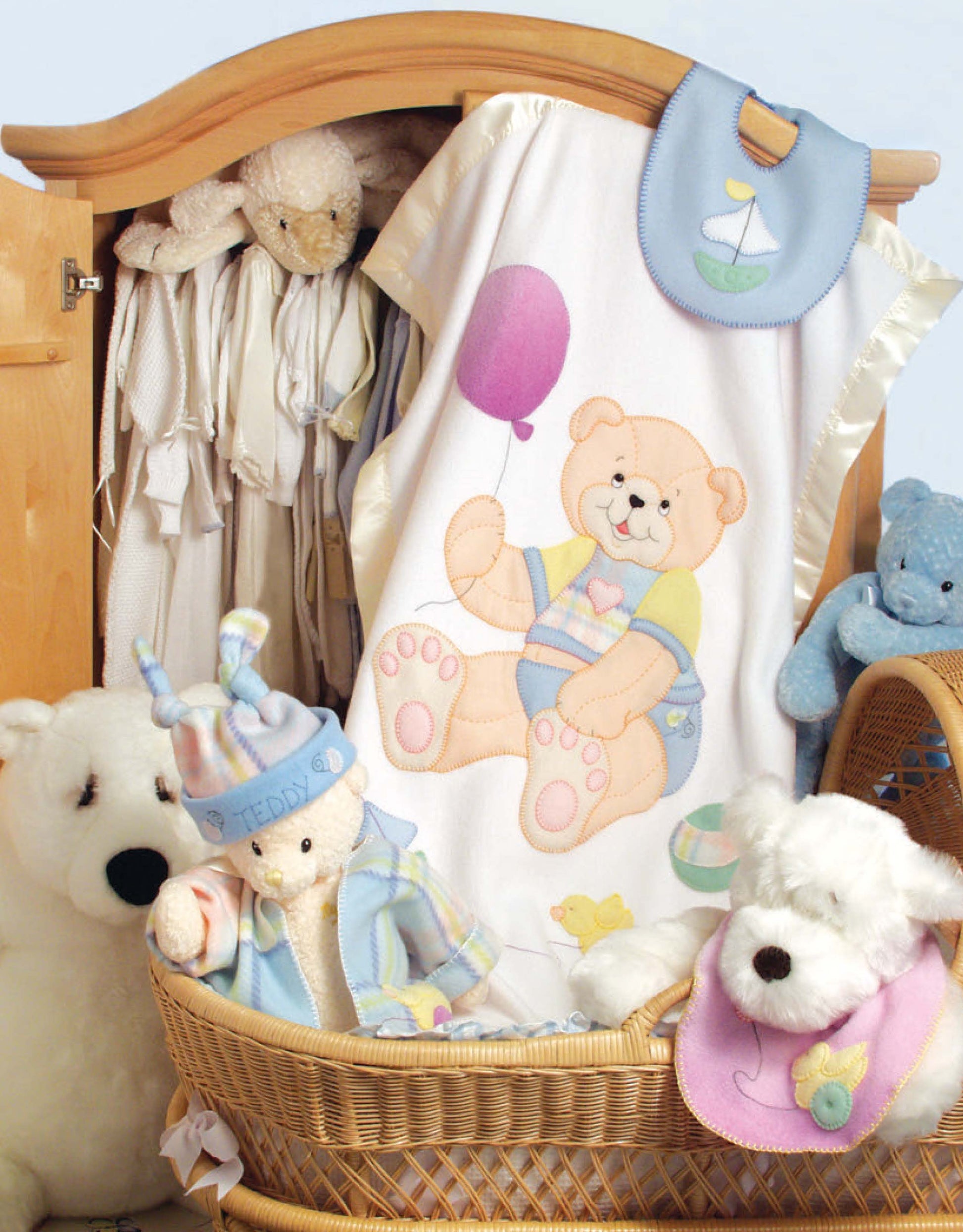 Pastel stuffed animals surround a basket with a white baby blanket featuring a cartoon bear, from "Baby Quilts and More—18 Projects" by Leisure Arts, beside a wooden wardrobe of hanging baby clothes.