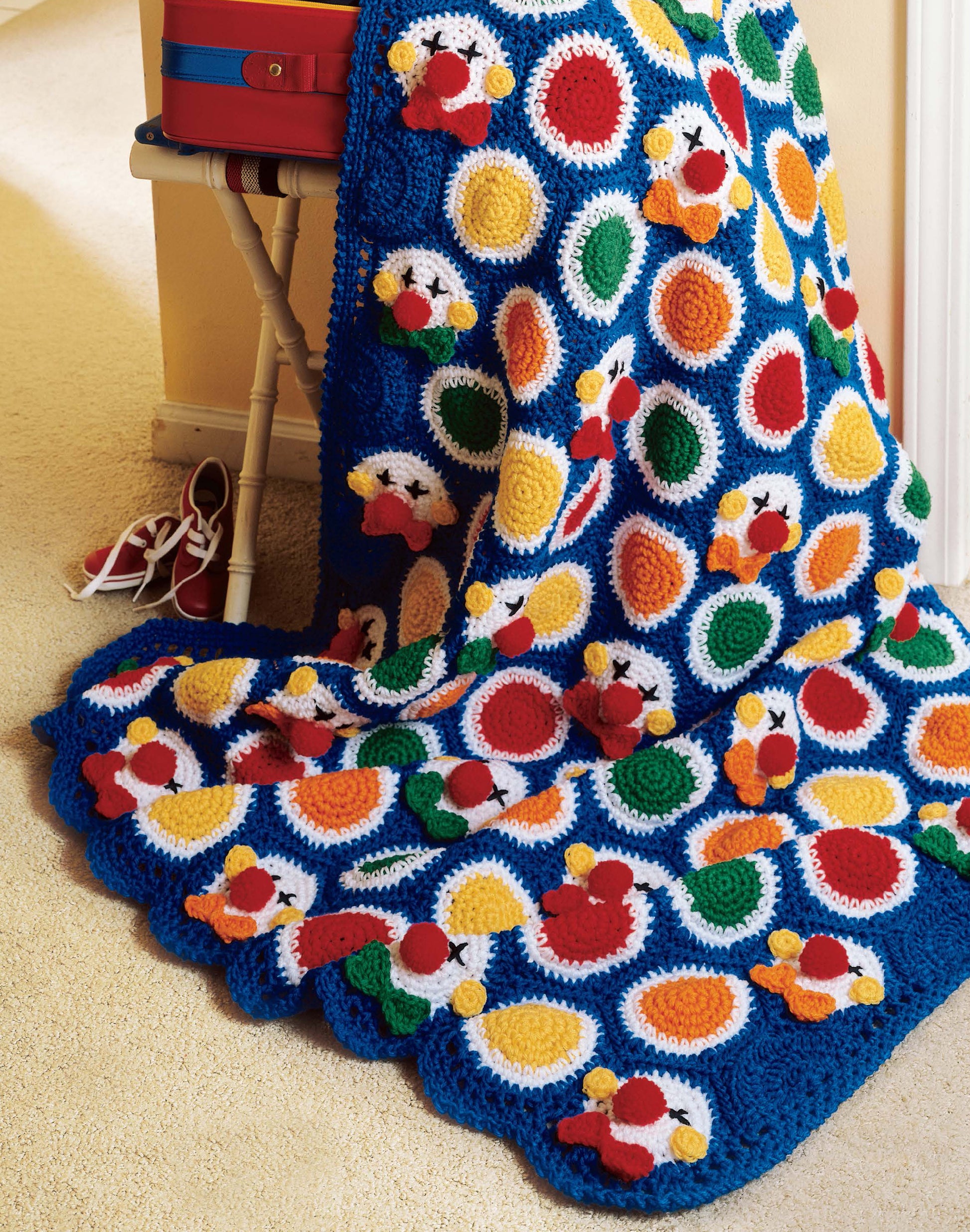 The Afghans by the Alphabet blanket from Leisure Arts features blue edges and rows of bright red, yellow, green, and orange circles. It's shown draped over a white bench with red shoes and a suitcase on a beige carpet.