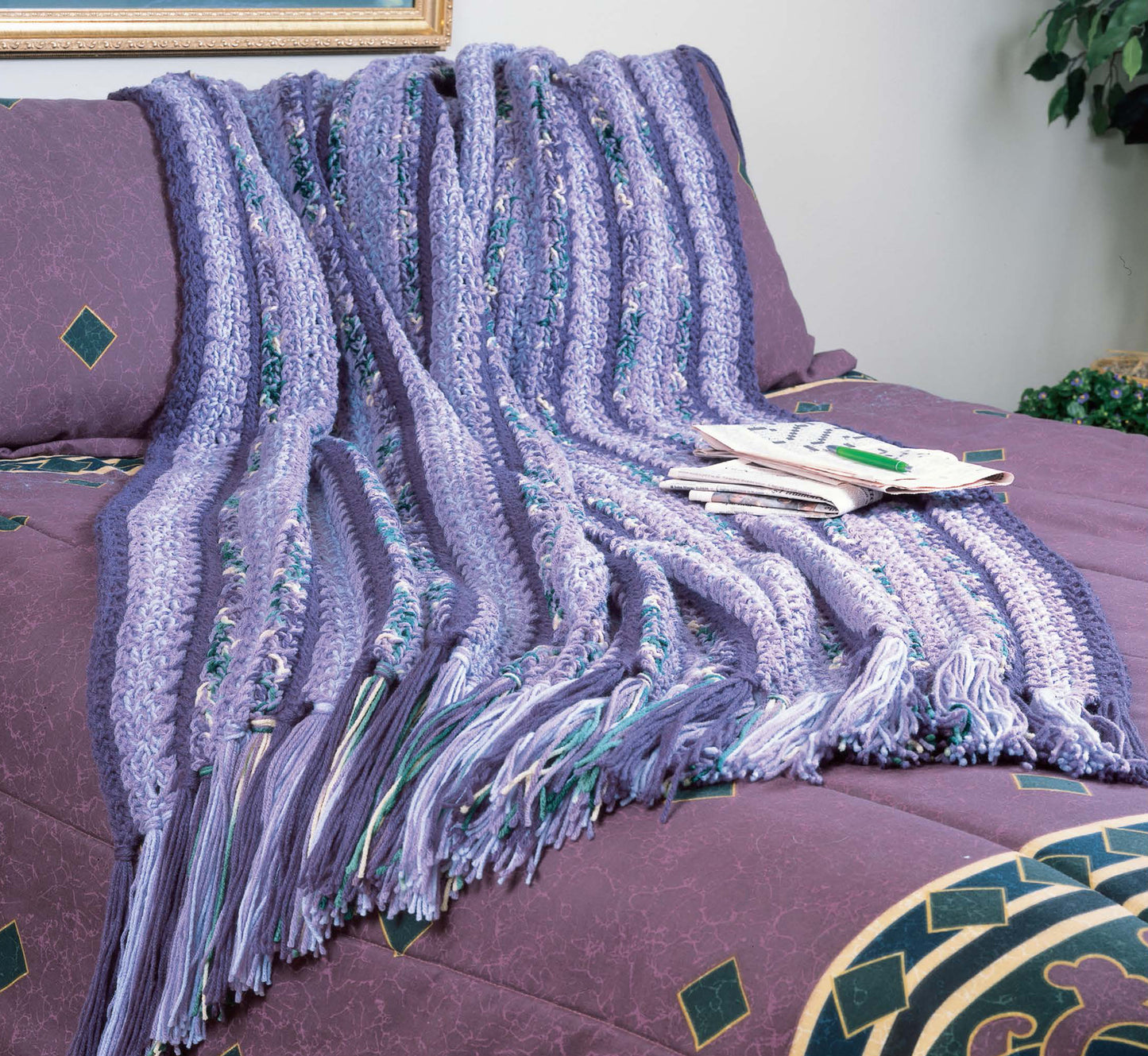 A purple, blue, and white striped crochet afghan from "Afghans by the Alphabet" by Leisure Arts lies on a purple-patterned bed. With magazines nearby, this cozy afghan is a perfect thoughtful crochet gift idea.