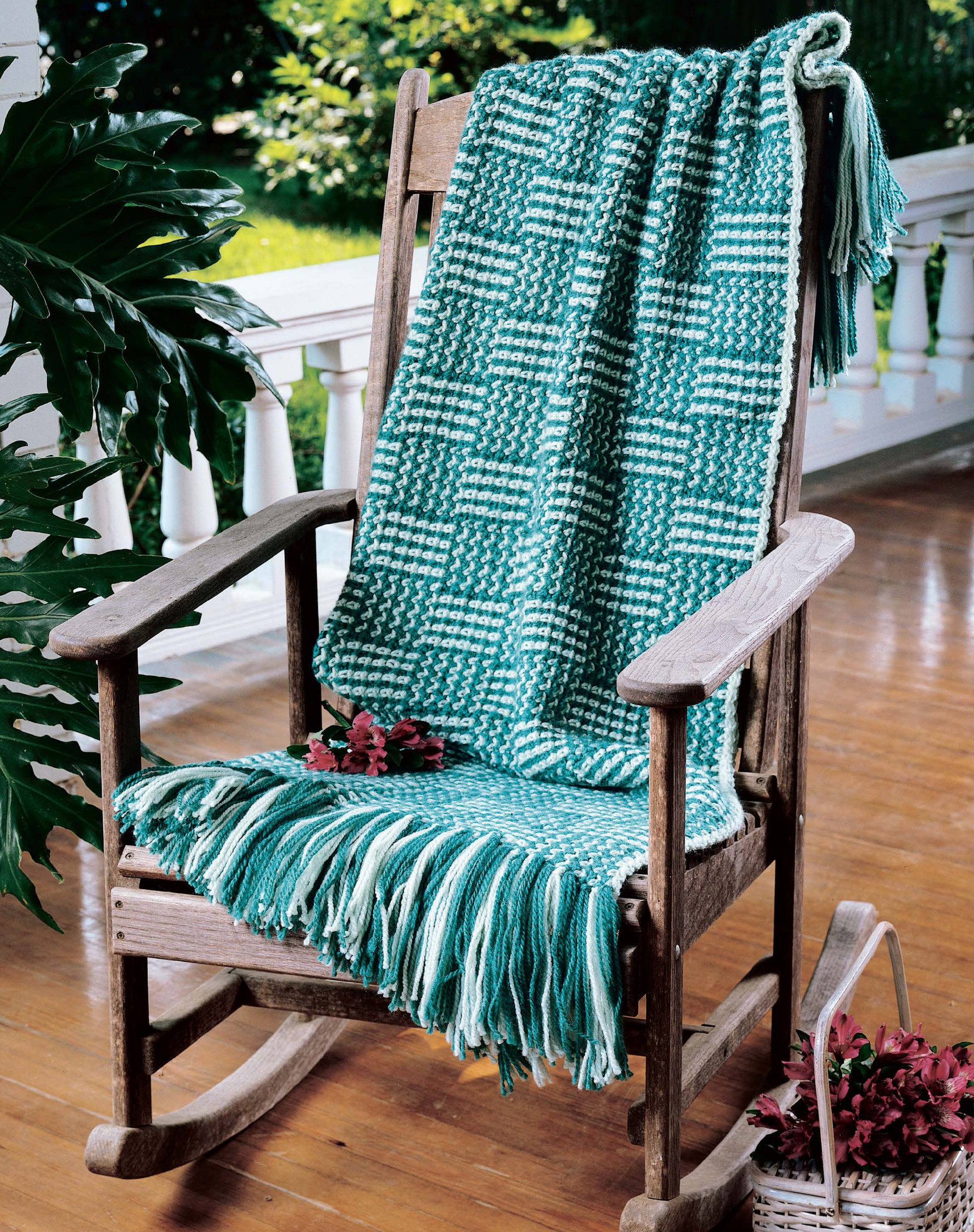 A wooden rocking chair with a green and white blanket on a sunlit porch creates the perfect cozy setting, inspiring crochet ideas from "Afghans by the Alphabet" by Leisure Arts.