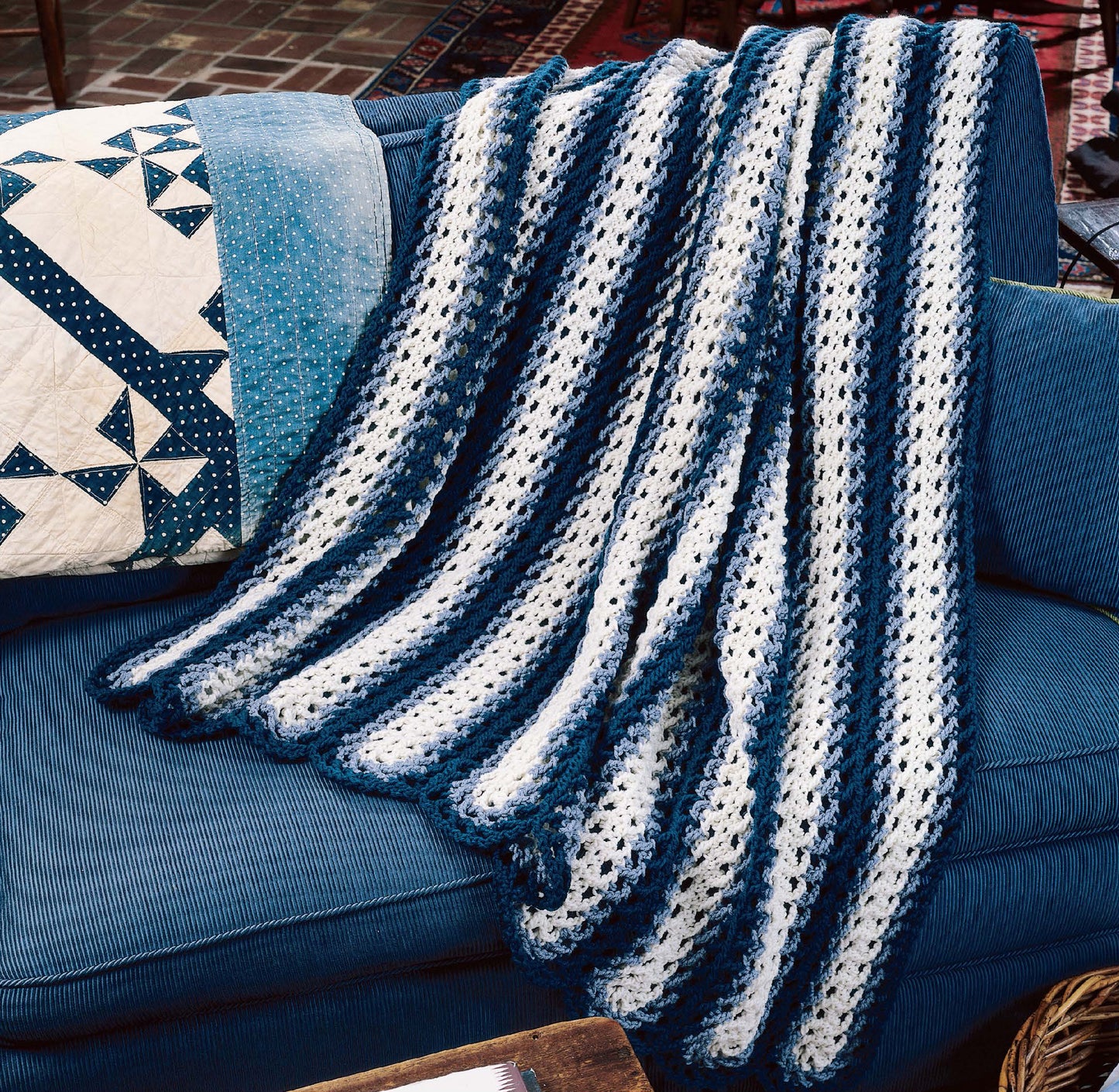 A cozy “Afghans by the Alphabet” crocheted blanket from Leisure Arts in blue and white stripes is draped over a blue couch beside a decorative pillow with a blue and white patchwork quilt design.