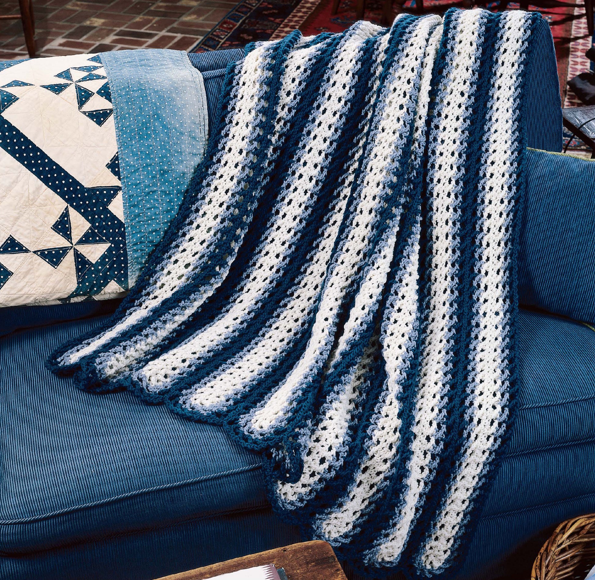 A cozy “Afghans by the Alphabet” crocheted blanket from Leisure Arts in blue and white stripes is draped over a blue couch beside a decorative pillow with a blue and white patchwork quilt design.