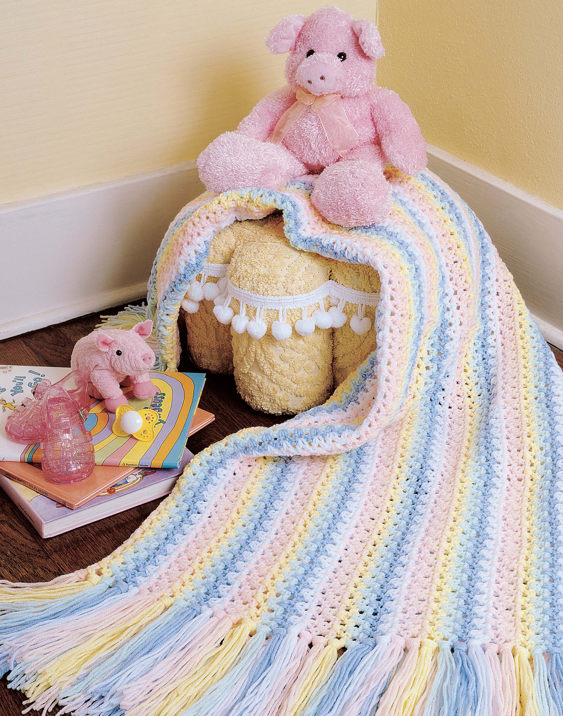The Leisure Arts "Afghans by the Alphabet" pastel striped crochet blanket with fringe covers a small stool, topped with two pink stuffed animals, children's books, and a pink toy on a dark floor beside a yellow wall.