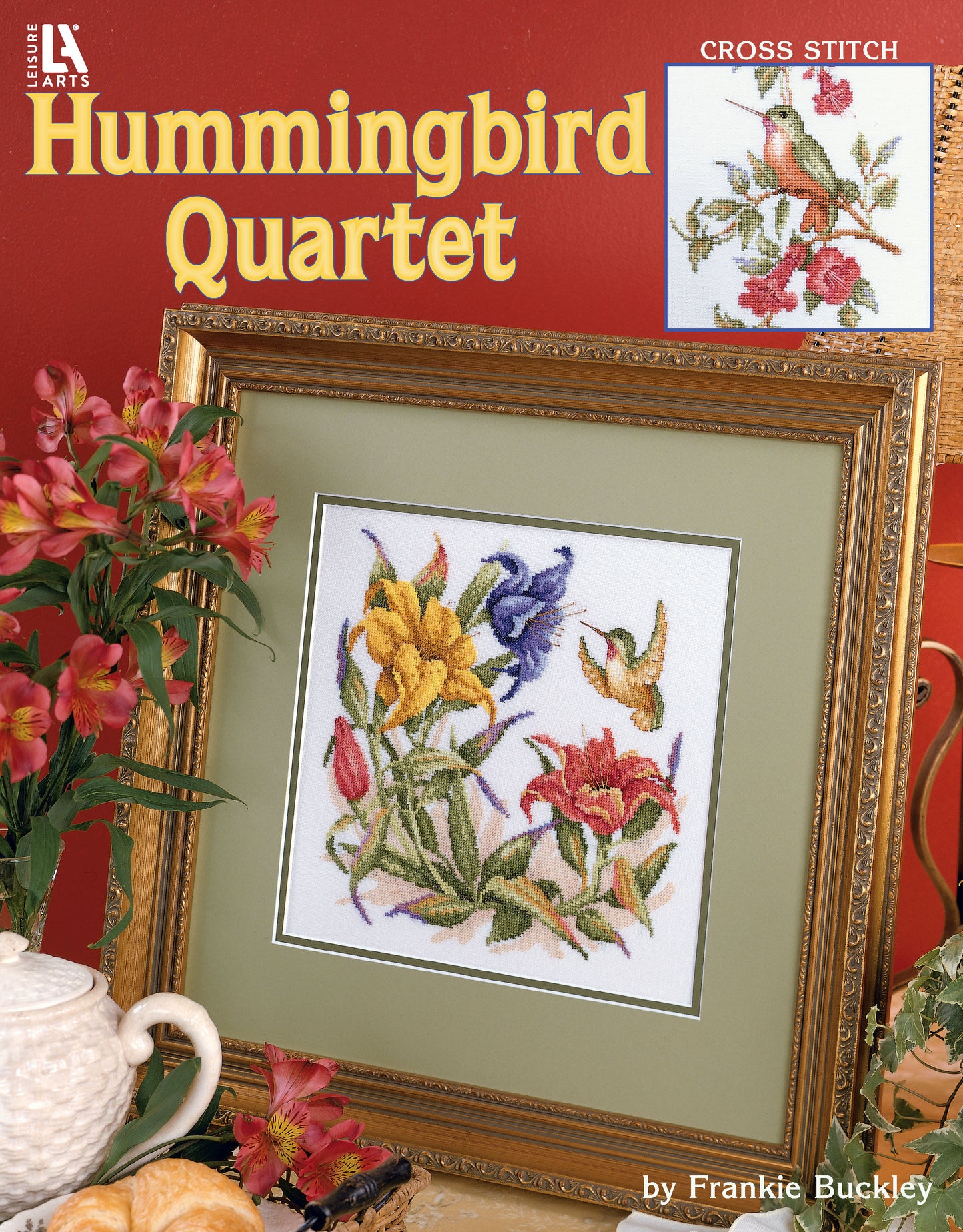 Framed cross-stitch "Hummingbird Quartet" by Leisure Arts features colorful hummingbirds among flowers, displayed with pink lilies and a gold-framed mirror for added charm.