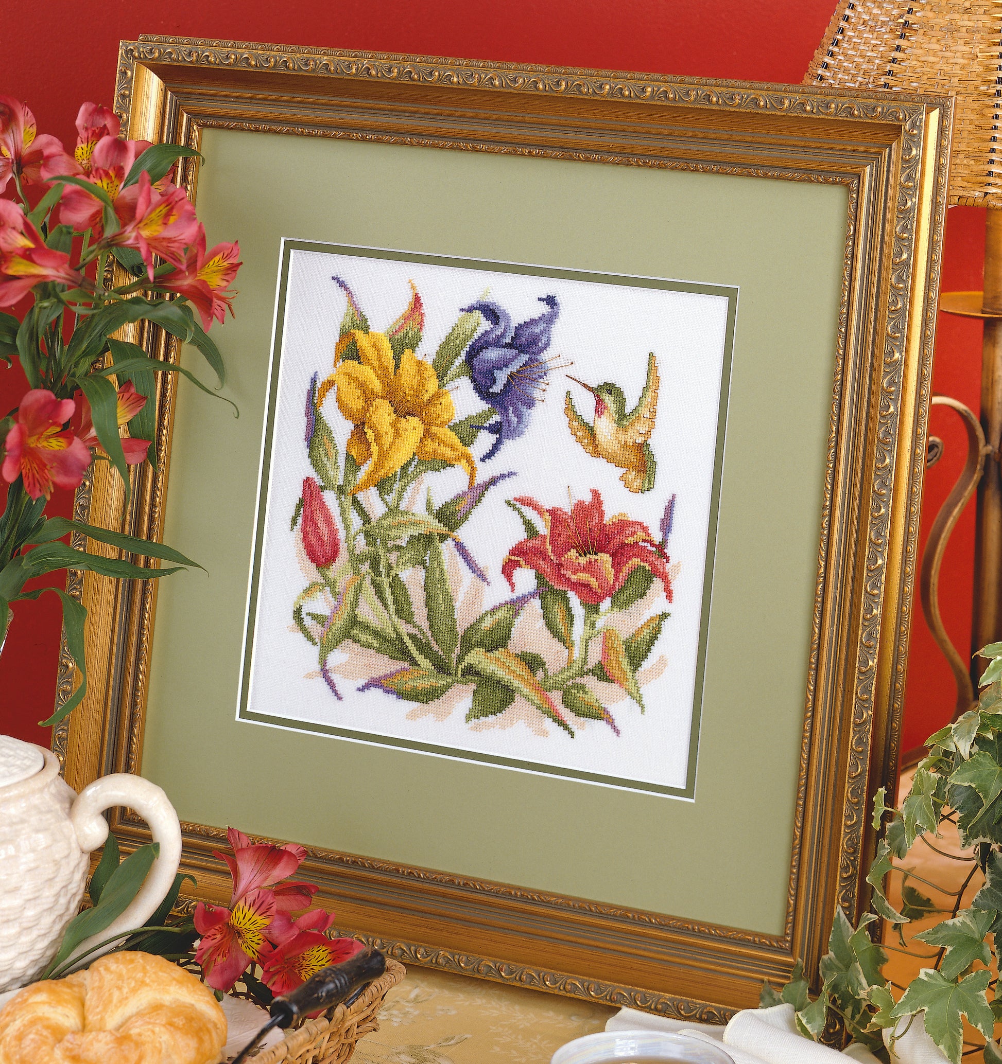 The Hummingbird Quartet Cross Stitch by Leisure Arts features colorful flowers and a hummingbird, displayed in a frame with fresh flowers, a teapot, and pastries on a table against a red wall. Pattern available from Leisure Arts.