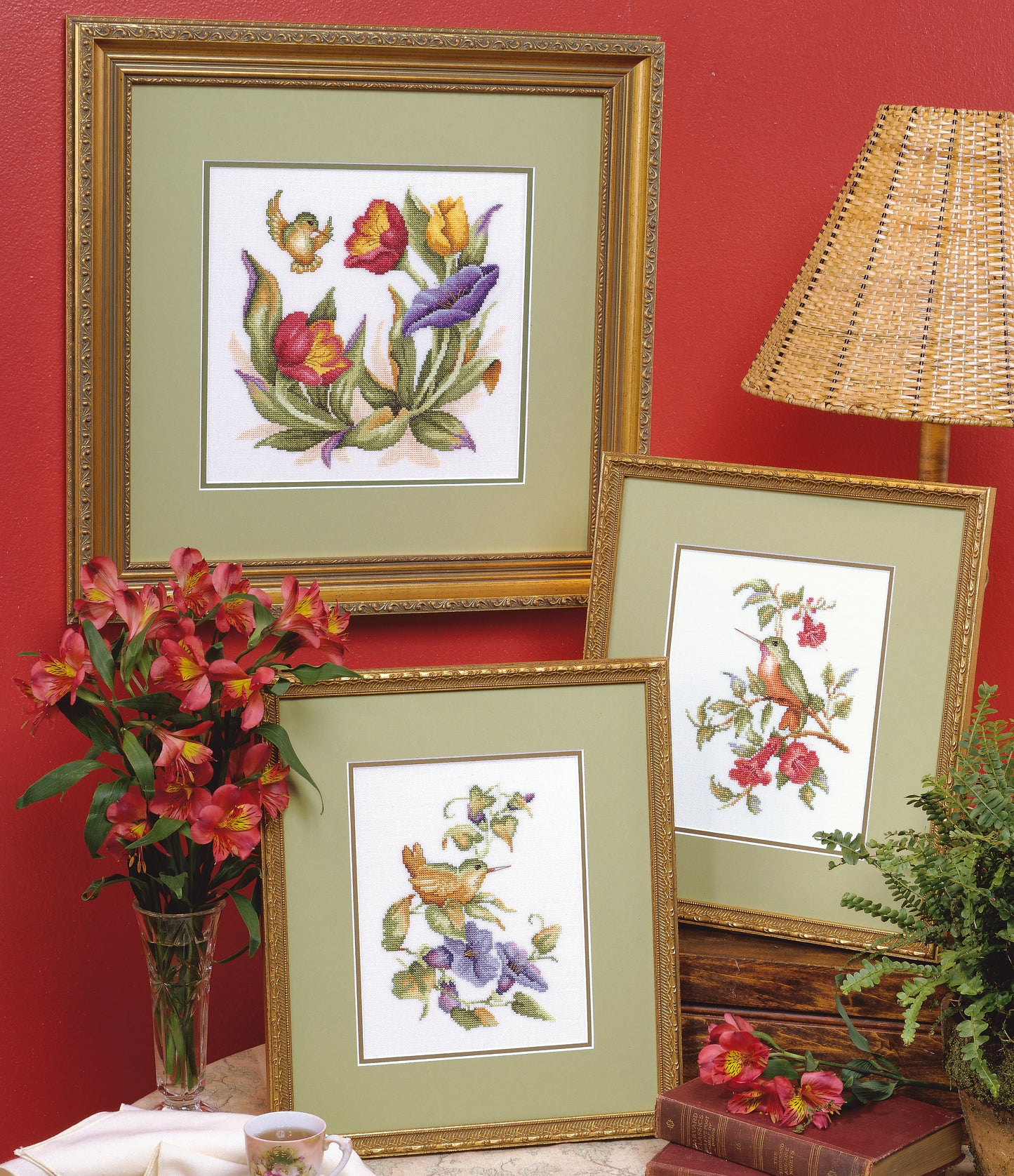 Three framed Hummingbird Quartet Cross Stitch artworks by Leisure Arts are arranged on a table against a red wall, beside a wicker lampshade, a vase of red flowers, and a green fern in this decorative scene.