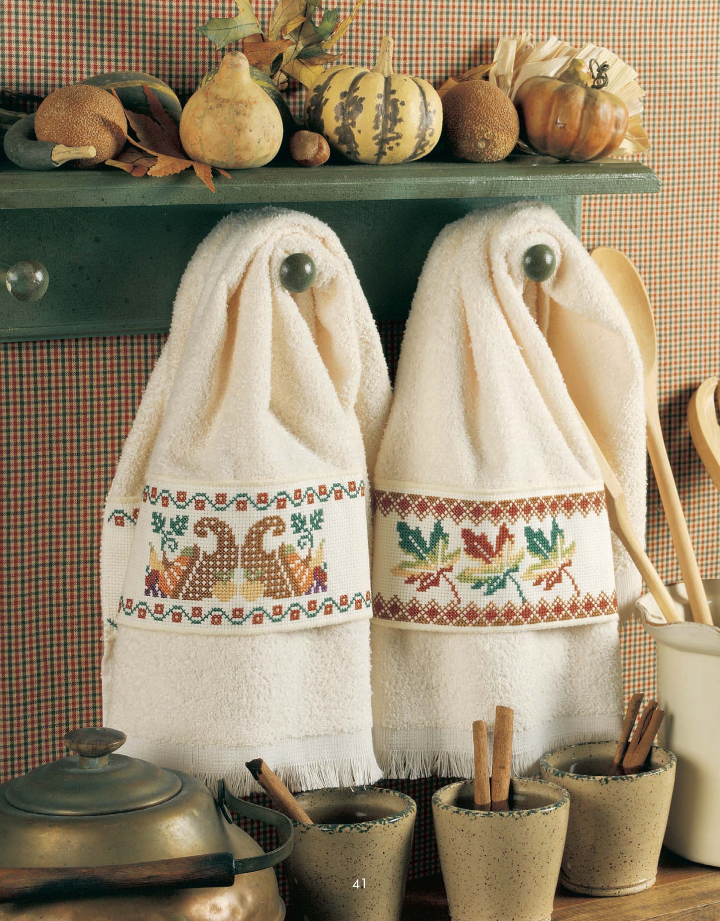 Two white towels stitched with Leisure Arts’ “Something for Every Season—56 Cross Stitch Designs” hang beneath a green shelf adorned with pumpkins, leaves, and gourds; pots and cinnamon sticks accent the festive scene below.