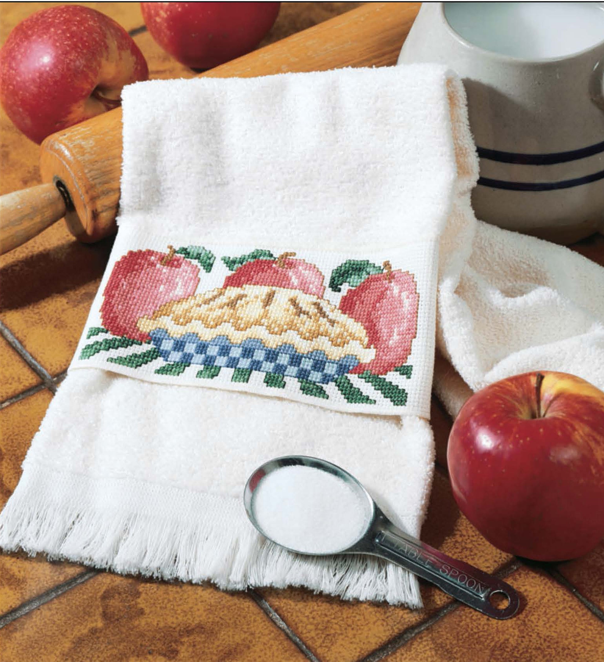 The Leisure Arts "Something for Every Season—56 Cross Stitch Designs" digital download features cross stitch patterns like apple pie and apples, shown here on a white towel alongside red apples, sugar, a rolling pin, and a ceramic bowl.