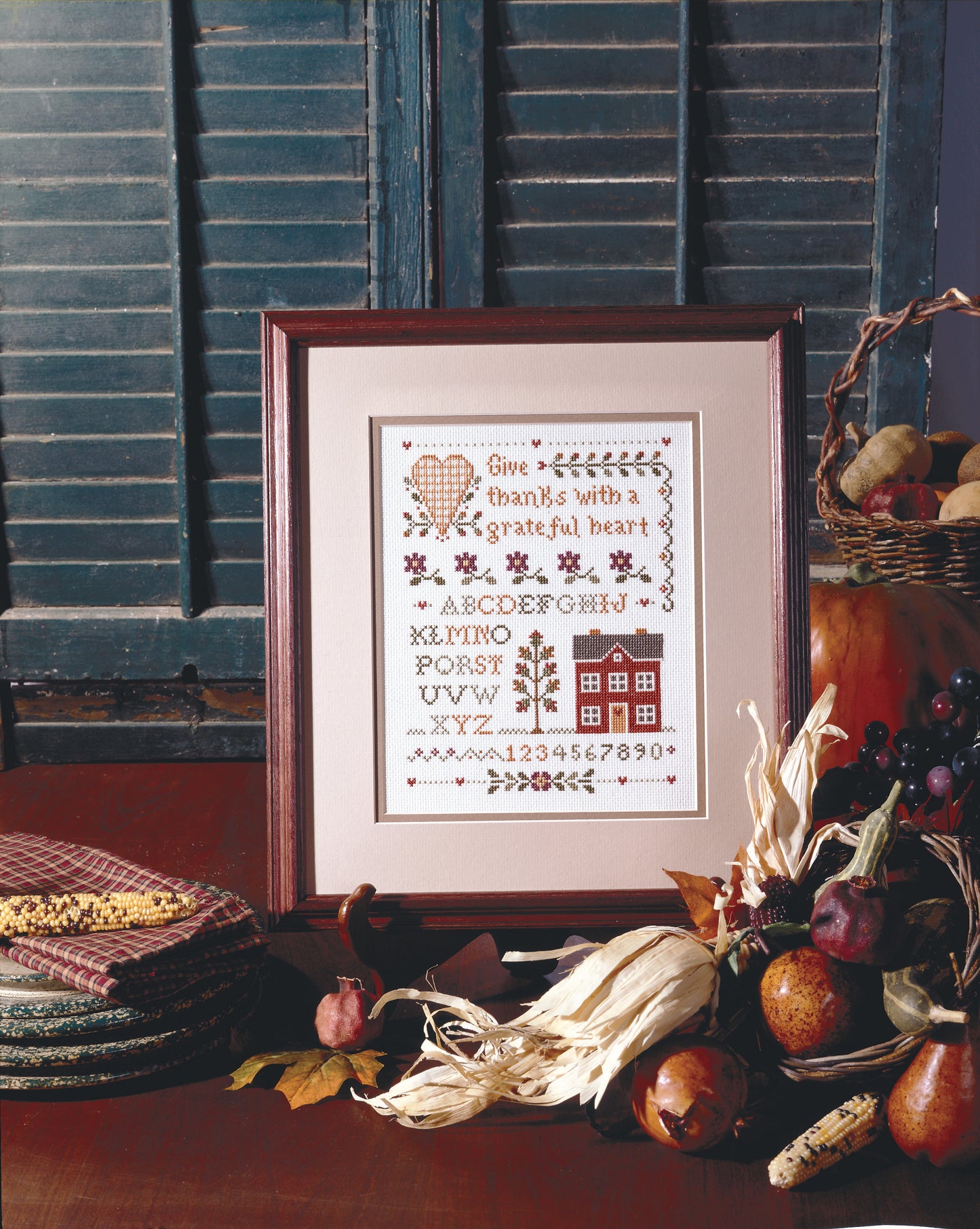 The Leisure Arts "A Sampling of Tradition Cross Stitch" is framed and displayed on a table with dried corn, pumpkins, grapes, apples, and stacked fabric before blue shutters—a charming autumn scene.