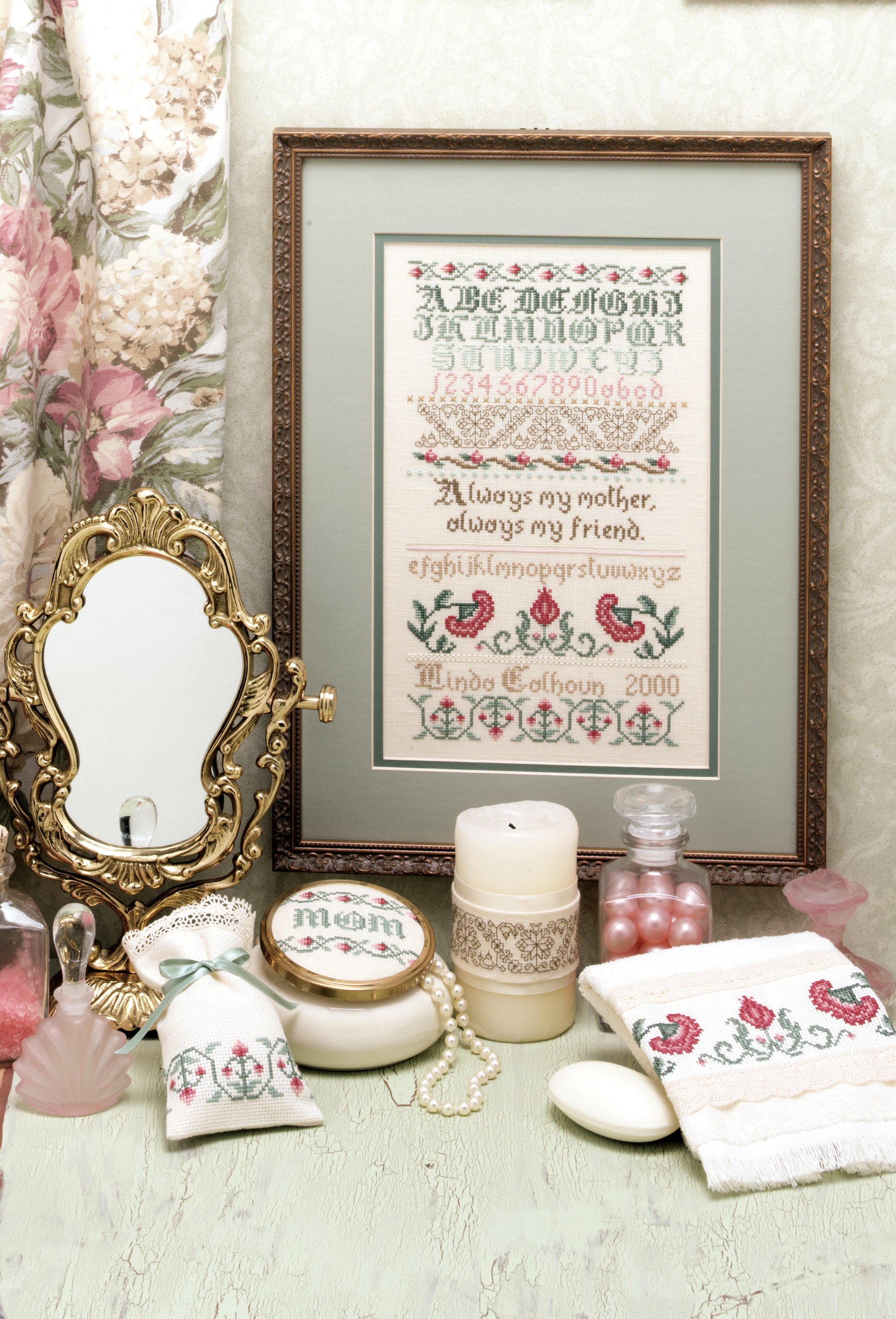 A decorative vanity with vintage perfume bottles, embroidered linens, a candle, and the Leisure Arts "A Sampling of Tradition Cross Stitch," featuring an alphabet, floral motifs, and the phrase "Always my mother, always my friend.
