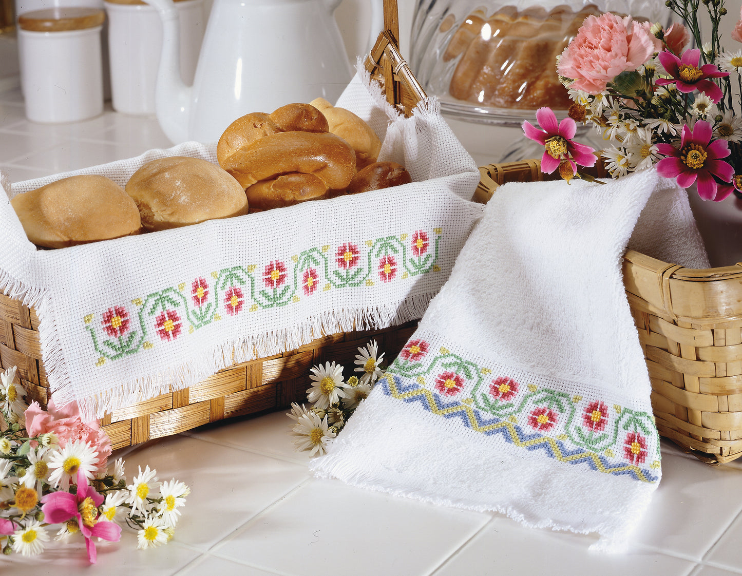 On a white countertop, bread rolls rest in a basket lined with Leisure Arts' A Sampling of Tradition Cross Stitch towel, beside another woven basket with flowers and a matching embroidered towel draped over it.