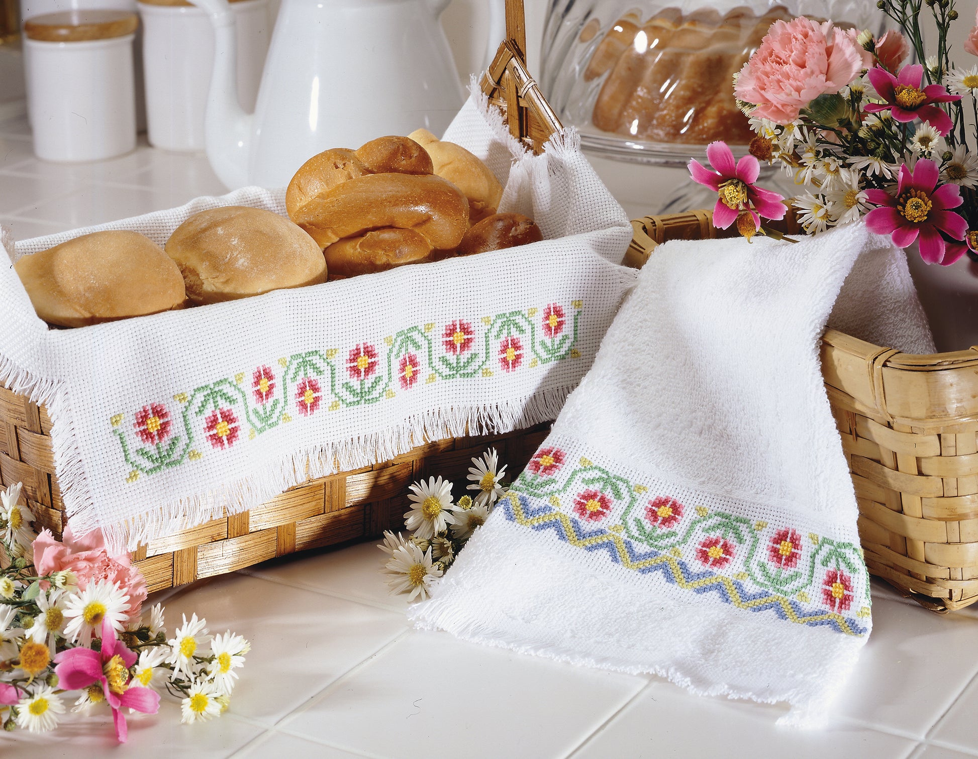 On a white countertop, bread rolls rest in a basket lined with Leisure Arts' A Sampling of Tradition Cross Stitch towel, beside another woven basket with flowers and a matching embroidered towel draped over it.