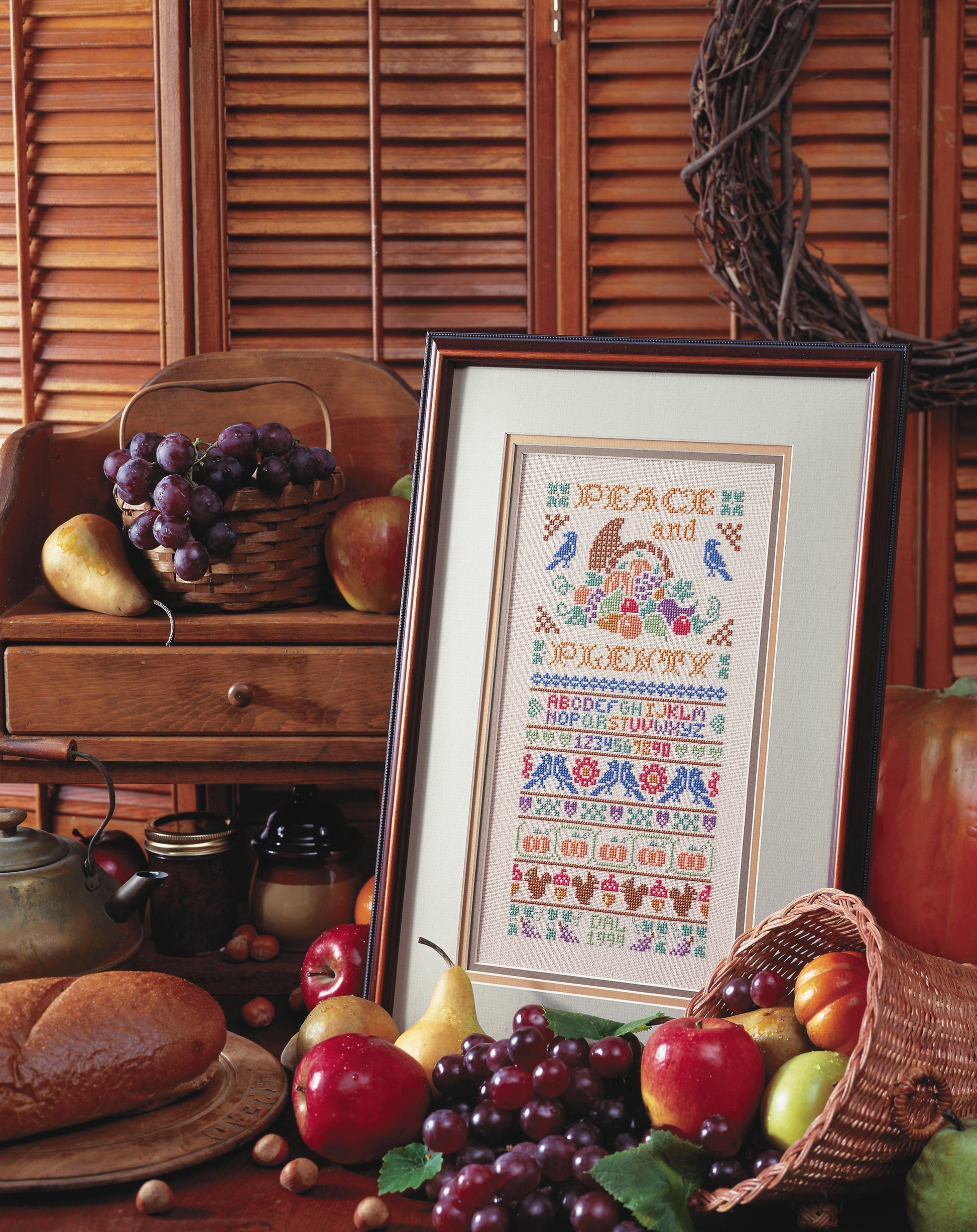 The Leisure Arts "A Sampling of Tradition Cross Stitch" features a framed design reading Peace and Plenty, displayed with fruit, bread, a teapot, and nuts—ideal for cozy stitchery or charming wall décor.