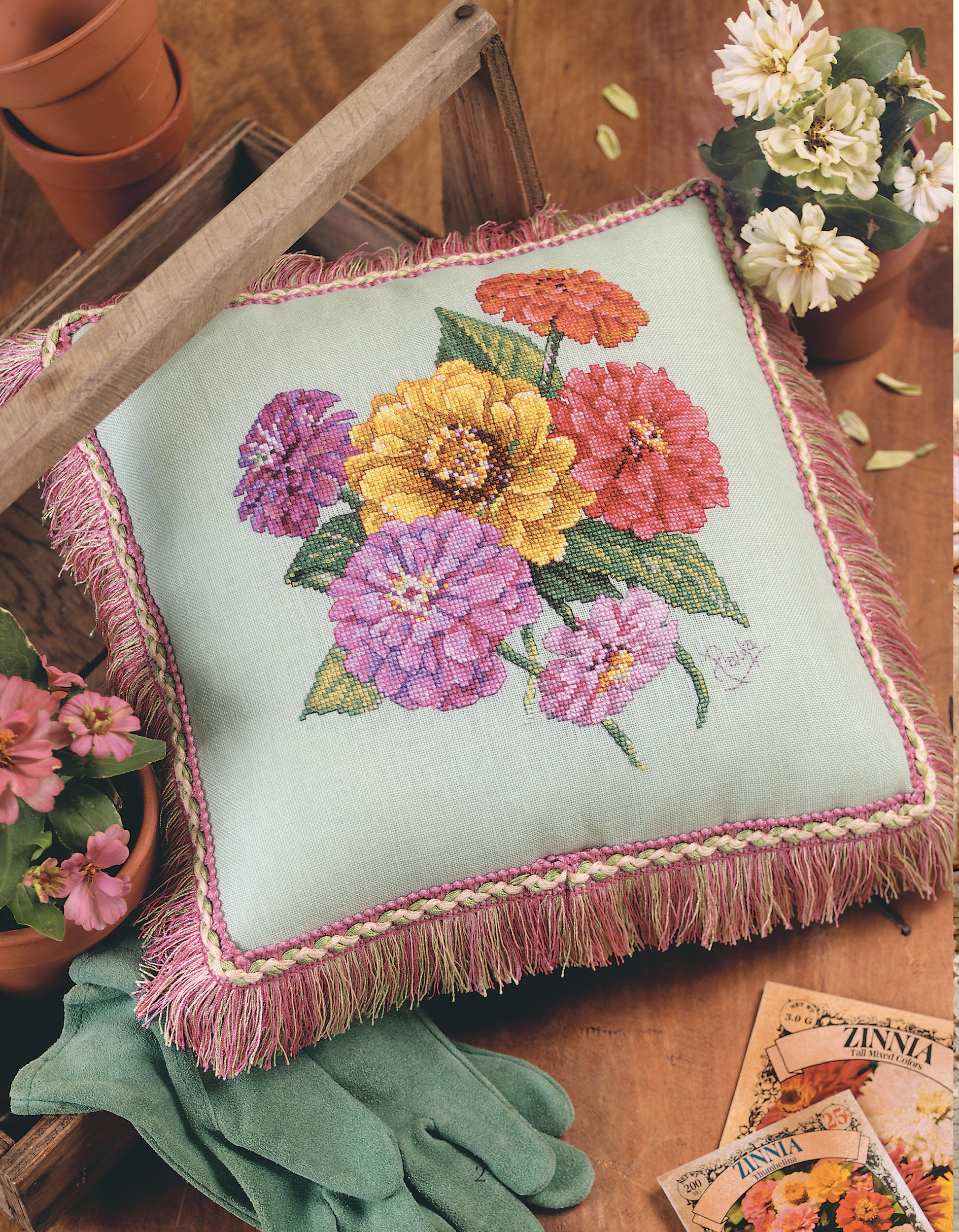 The "Pillows from Paula's Garden" by Leisure Arts, featuring embroidered pink, yellow, orange, and purple zinnias, rests on a wooden table with potted flowers, gloves, and seed packets.