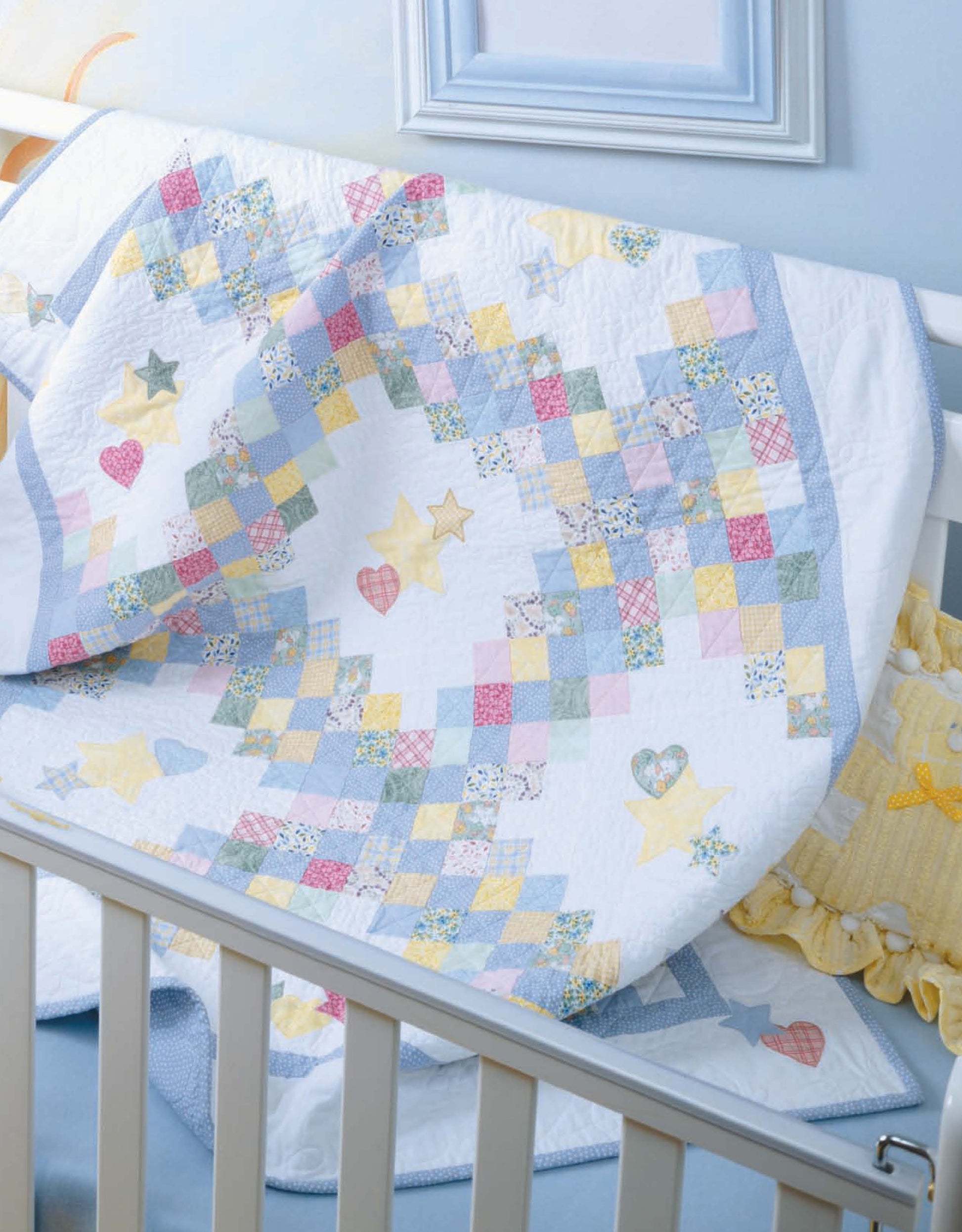 The Tadpole Quilts for Baby by Leisure Arts, featuring pastel squares, stars, and hearts with charming patterns, drapes over a white crib. Inside is a yellow ruffled pillow and above hangs a blue picture frame.