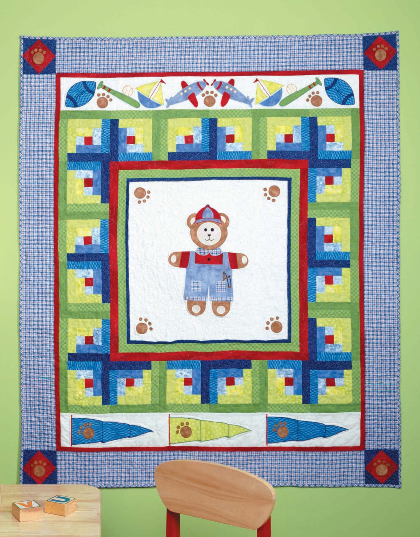 The Tadpole Quilts for Baby by Leisure Arts is a colorful patchwork quilt with a teddy bear center, featuring geometric shapes, sailboats, paw prints, and toys. It hangs on a light green wall above a wooden chair and blocks.