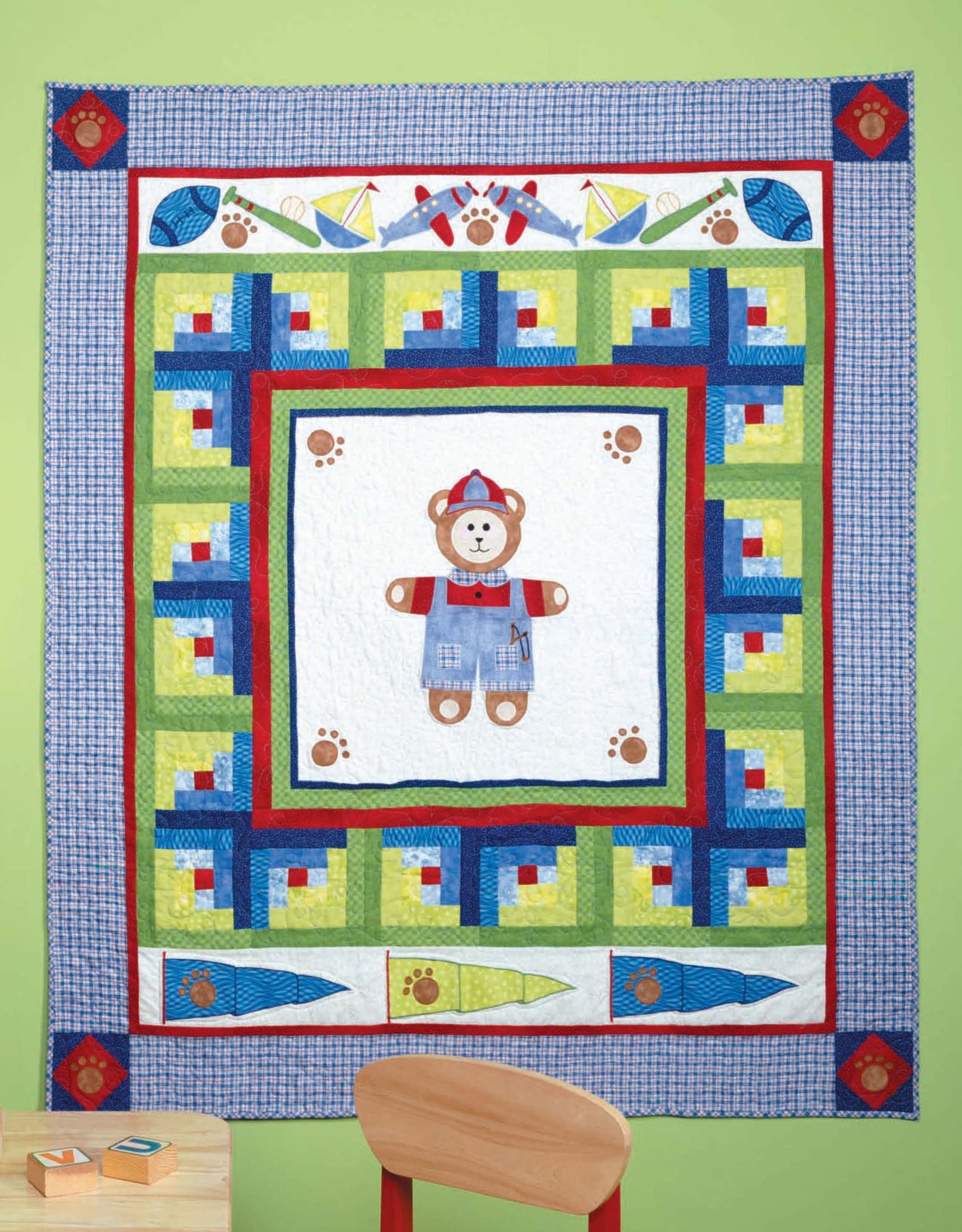 The Tadpole Quilts for Baby by Leisure Arts is a colorful patchwork quilt with a teddy bear center, featuring geometric shapes, sailboats, paw prints, and toys. It hangs on a light green wall above a wooden chair and blocks.