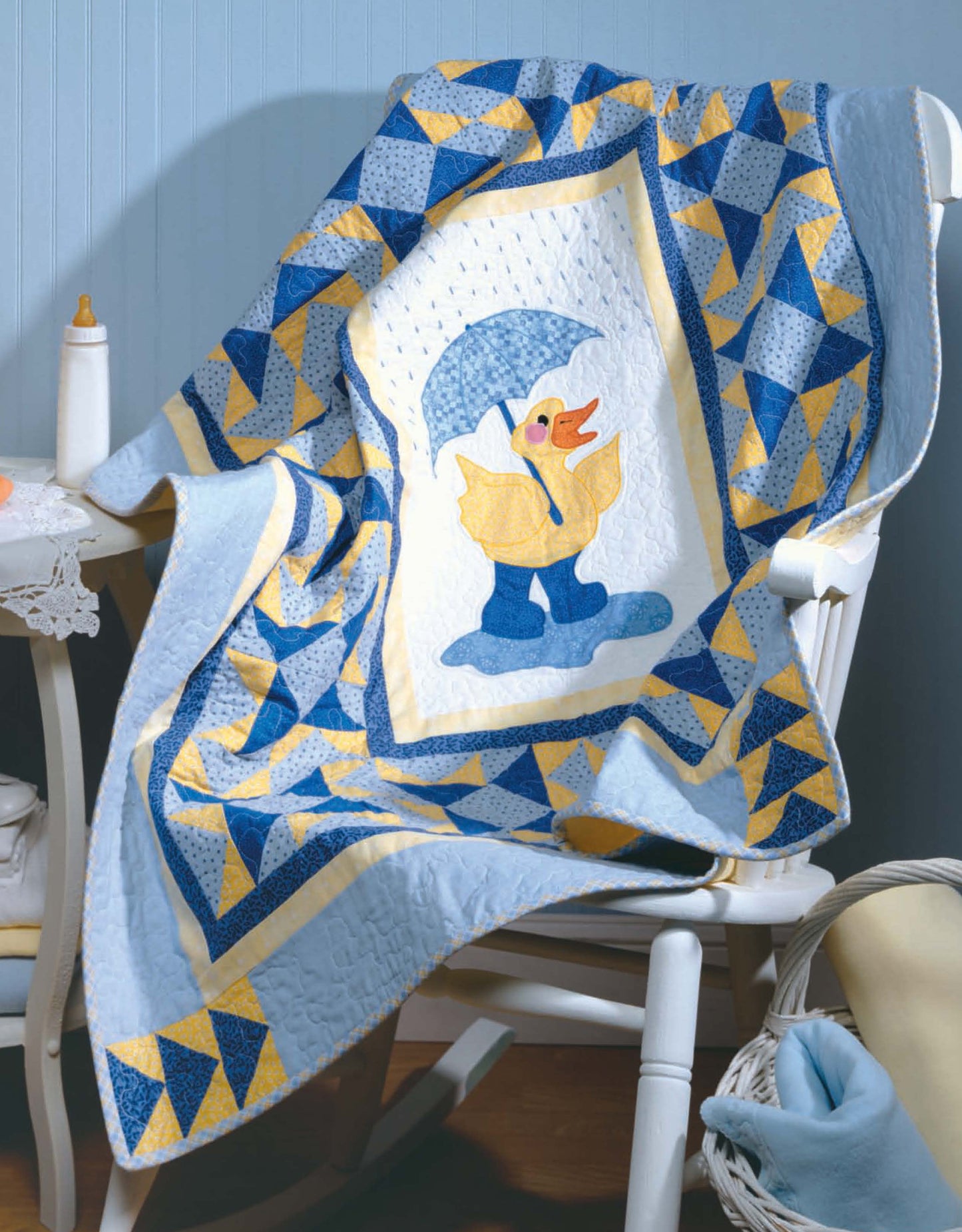 The Tadpole Quilts for Baby from Leisure Arts is a blue and yellow quilt with easy-to-piece blocks, featuring a central duck in rain boots and umbrella, shown draped over a white chair with a baby bottle and basket nearby.
