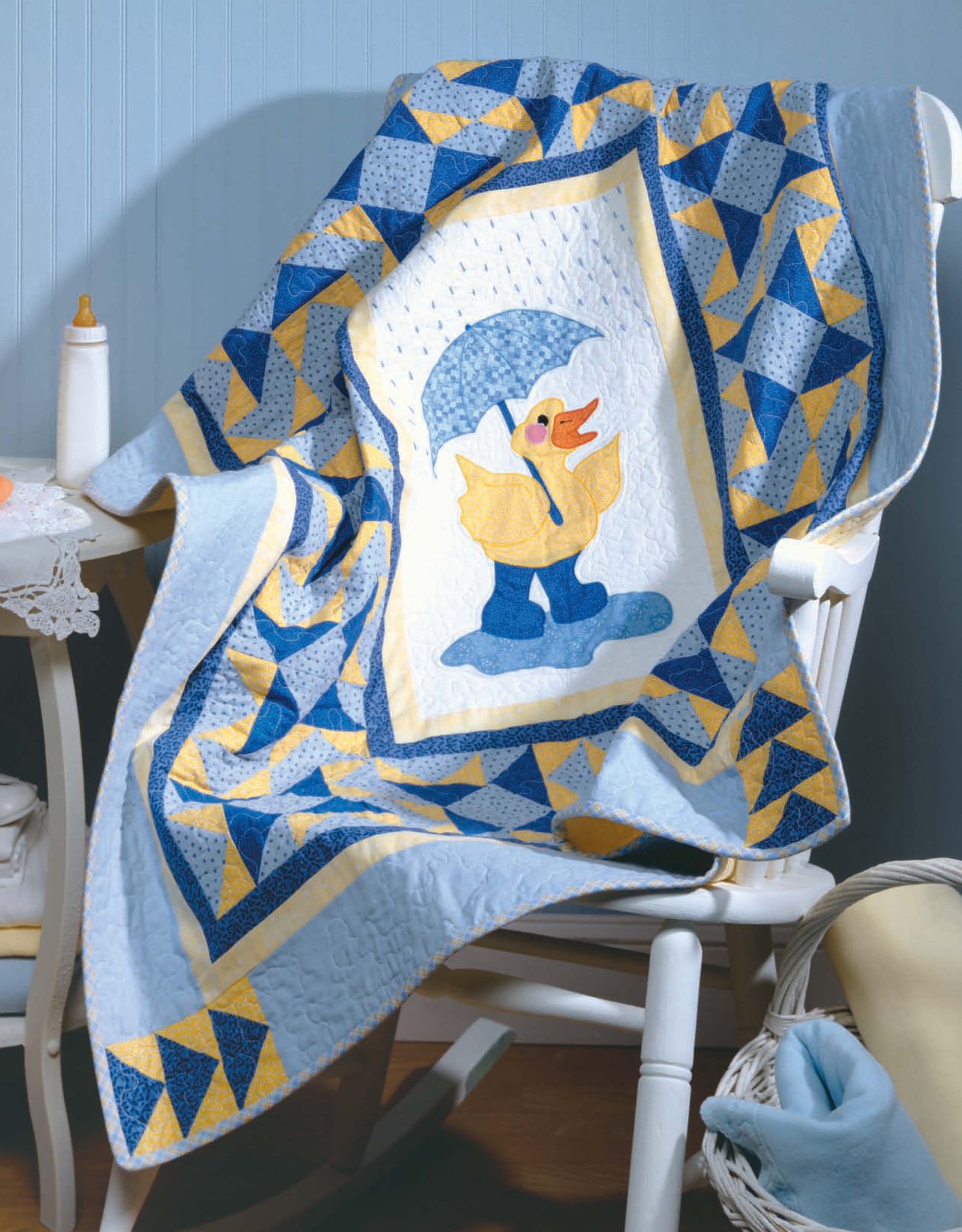 The Tadpole Quilts for Baby from Leisure Arts is a blue and yellow quilt with easy-to-piece blocks, featuring a central duck in rain boots and umbrella, shown draped over a white chair with a baby bottle and basket nearby.