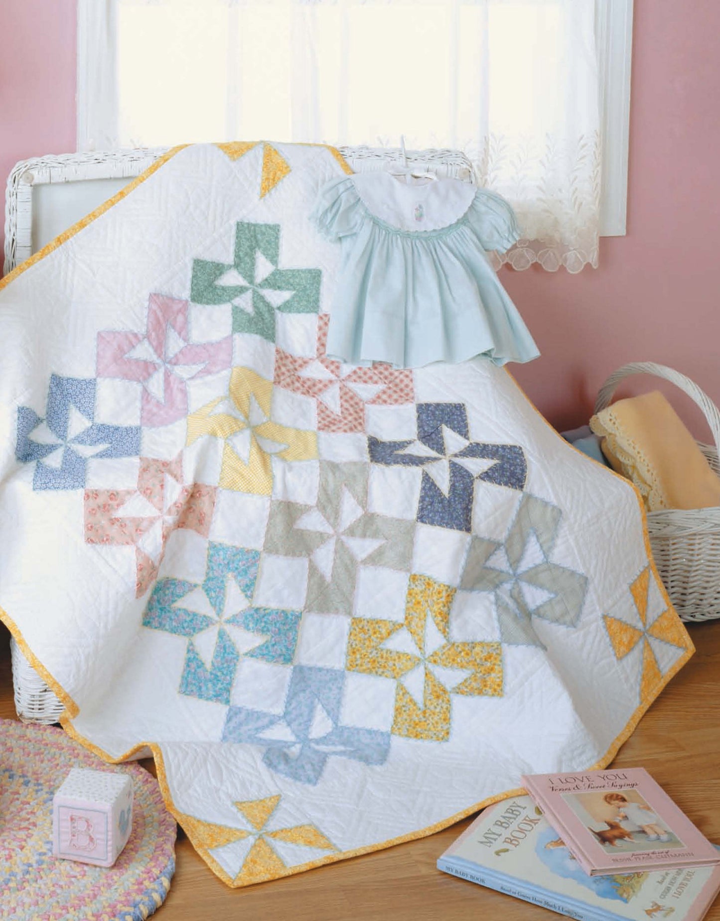 Leisure Arts’ Tadpole Quilts for Baby features a vibrant patchwork pinwheel quilt draped over a basket beside a blue baby dress, children’s books, and a toy block—perfect for adding cozy charm to nurseries.