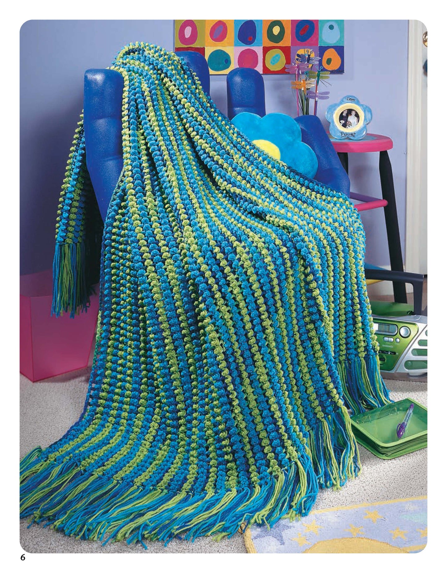 A vibrant blue and green striped throw from Color-Charged Throws by Leisure Arts, with easy crochet patterns and long fringe, covers a blue chair in a colorful room decorated with a flower pillow, clock, and polka dot artwork.
