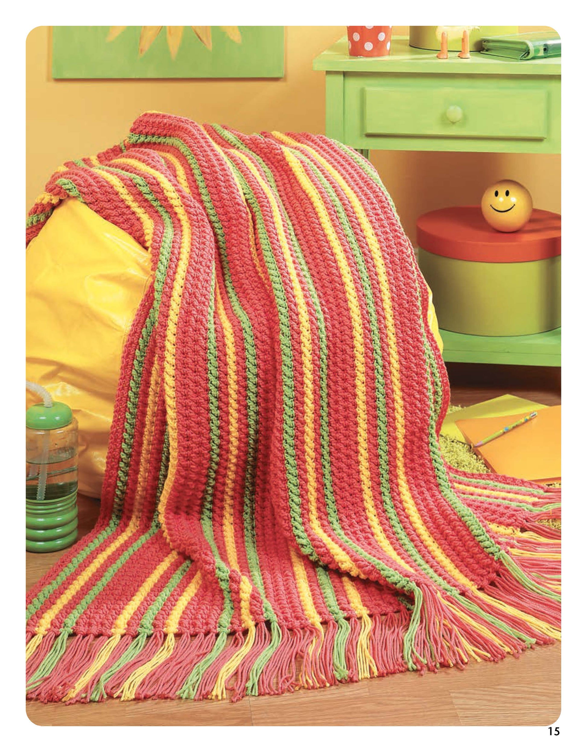The Leisure Arts Color-Charged Throws, a striped crochet blanket in pink, yellow, and green with fringed edges, is draped over a yellow bean bag in a cheerful room decorated with pastel furniture and playful accents.