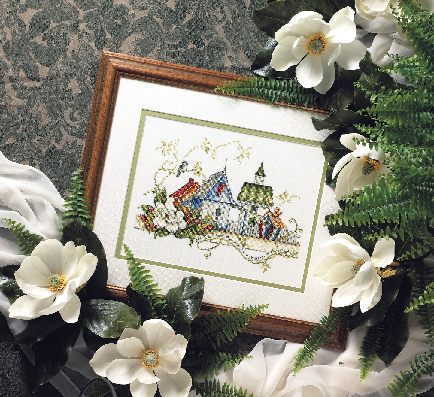 The Unity Birdhouses Cross Stitch by Leisure Arts is a framed artwork featuring birdhouses amid flowers and greenery, displayed with magnolia blossoms, green leaves, ferns, and white fabric on a patterned green background.