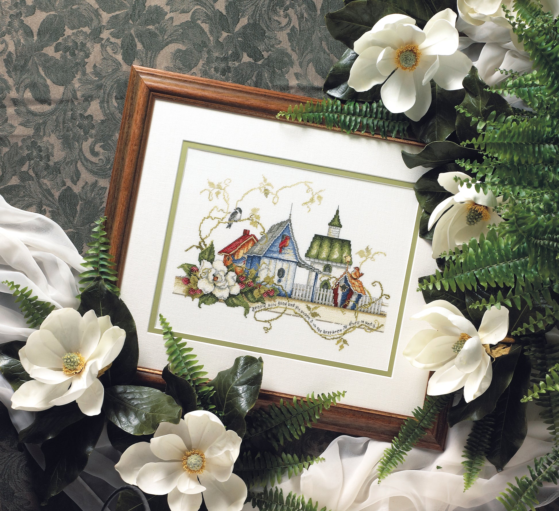 The Unity Birdhouses Cross Stitch by Leisure Arts is a framed artwork featuring birdhouses amid flowers and greenery, displayed with magnolia blossoms, green leaves, ferns, and white fabric on a patterned green background.