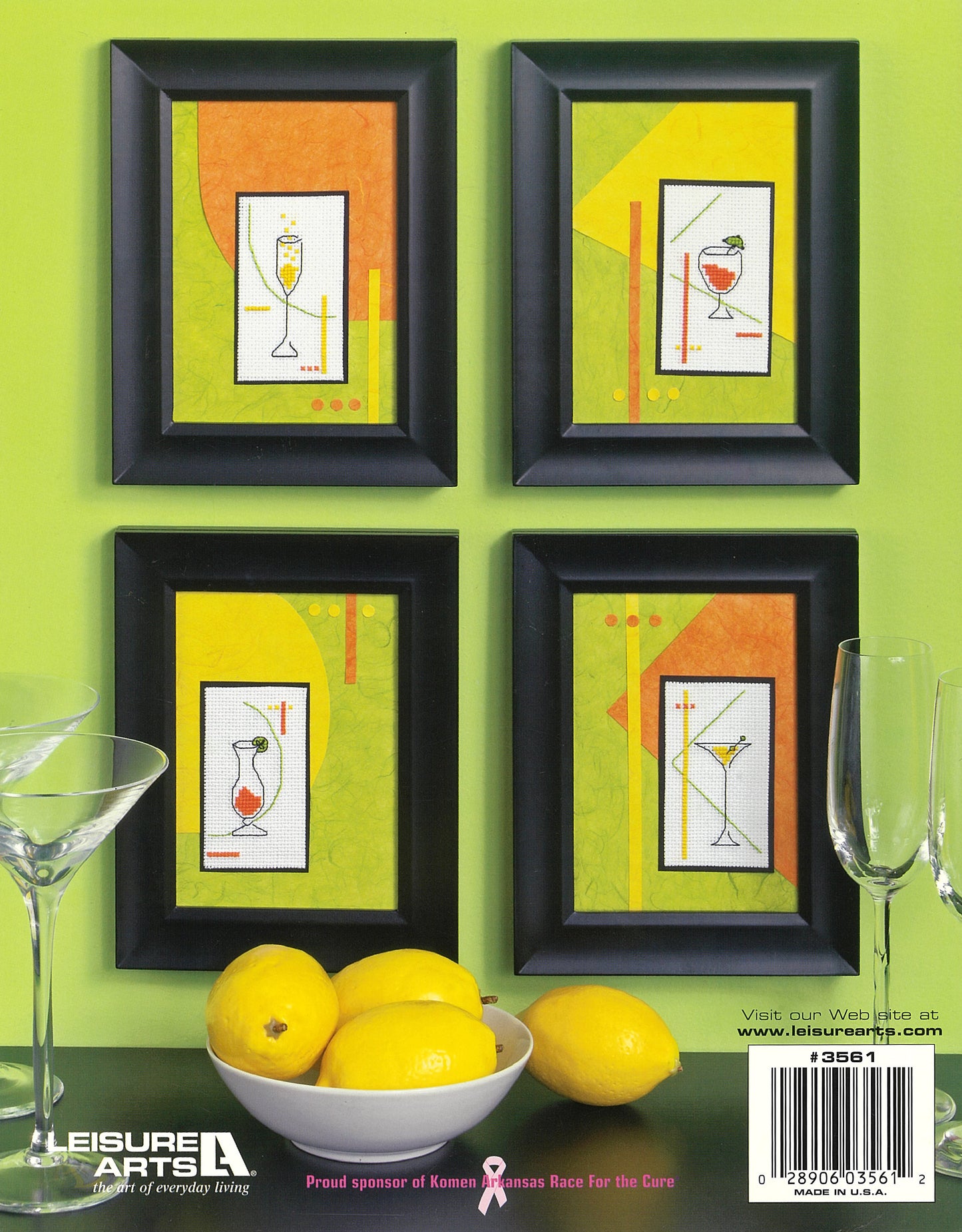 Four black-framed cocktail-themed cross stitch collages from Leisure Arts’ Quick & Easy Decorating Digital Download hang on a lime green wall above a table with three lemons, two empty wine glasses, and an instruction booklet.