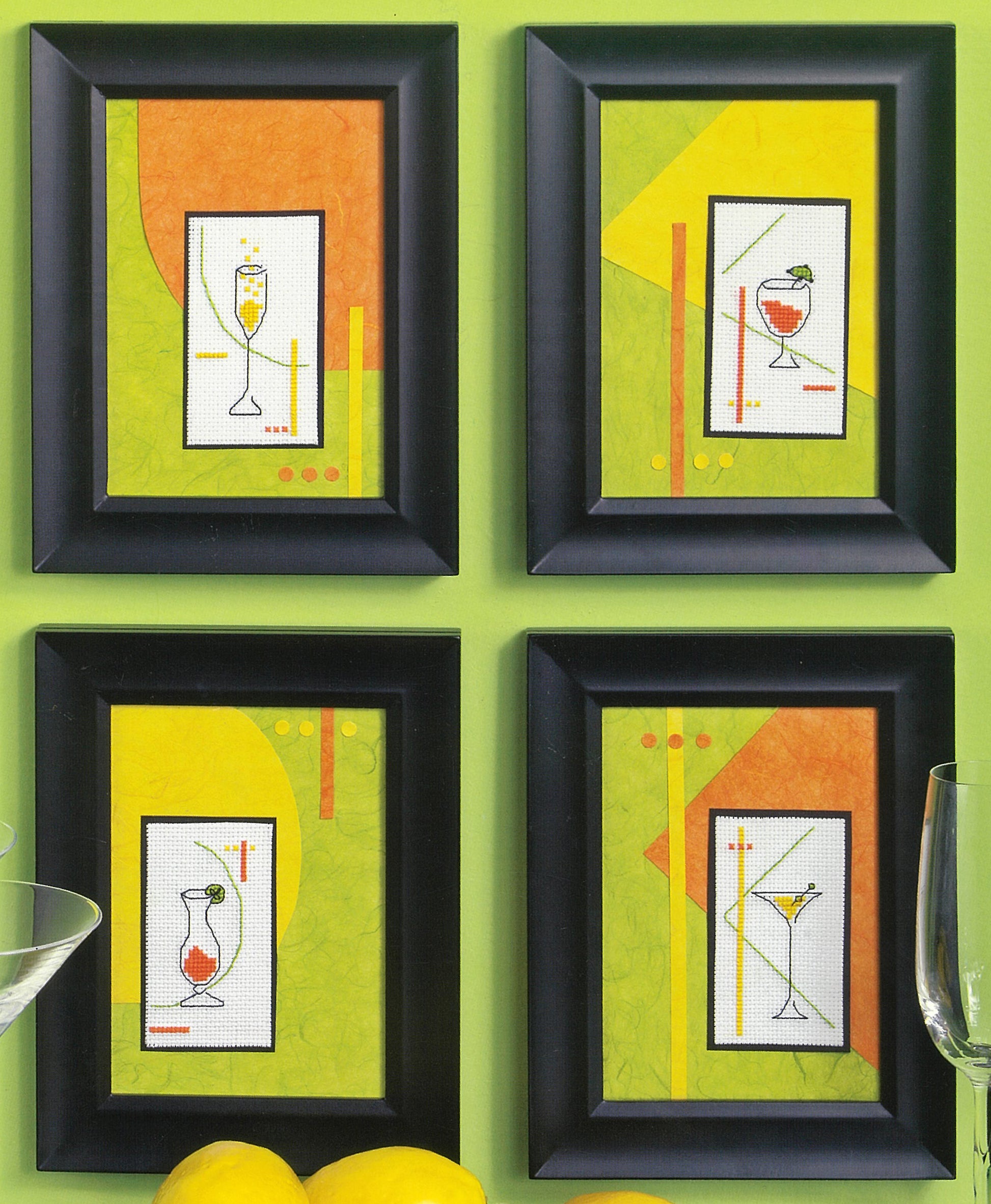 Four framed cocktail cross stitch collages from Leisure Arts' "Quick & Easy Decorating" hang on a green wall, each in a black frame with abstract backgrounds and unique drink designs. Lemons and cocktail glasses are seen at the bottom.