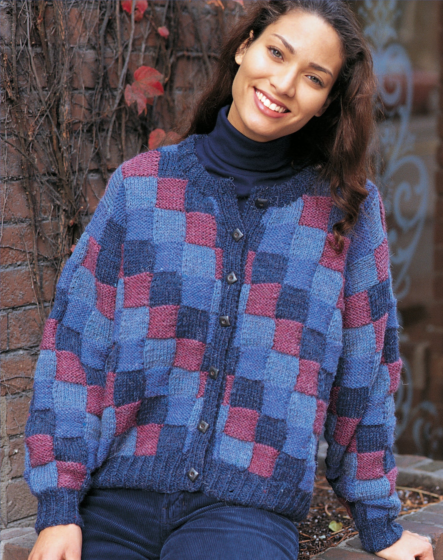 A woman smiles outdoors in The Sweater Collection Knit by Leisure Arts—a blue and purple checkered cardigan over a navy turtleneck and jeans—standing against a brick wall with vines and red leaves.