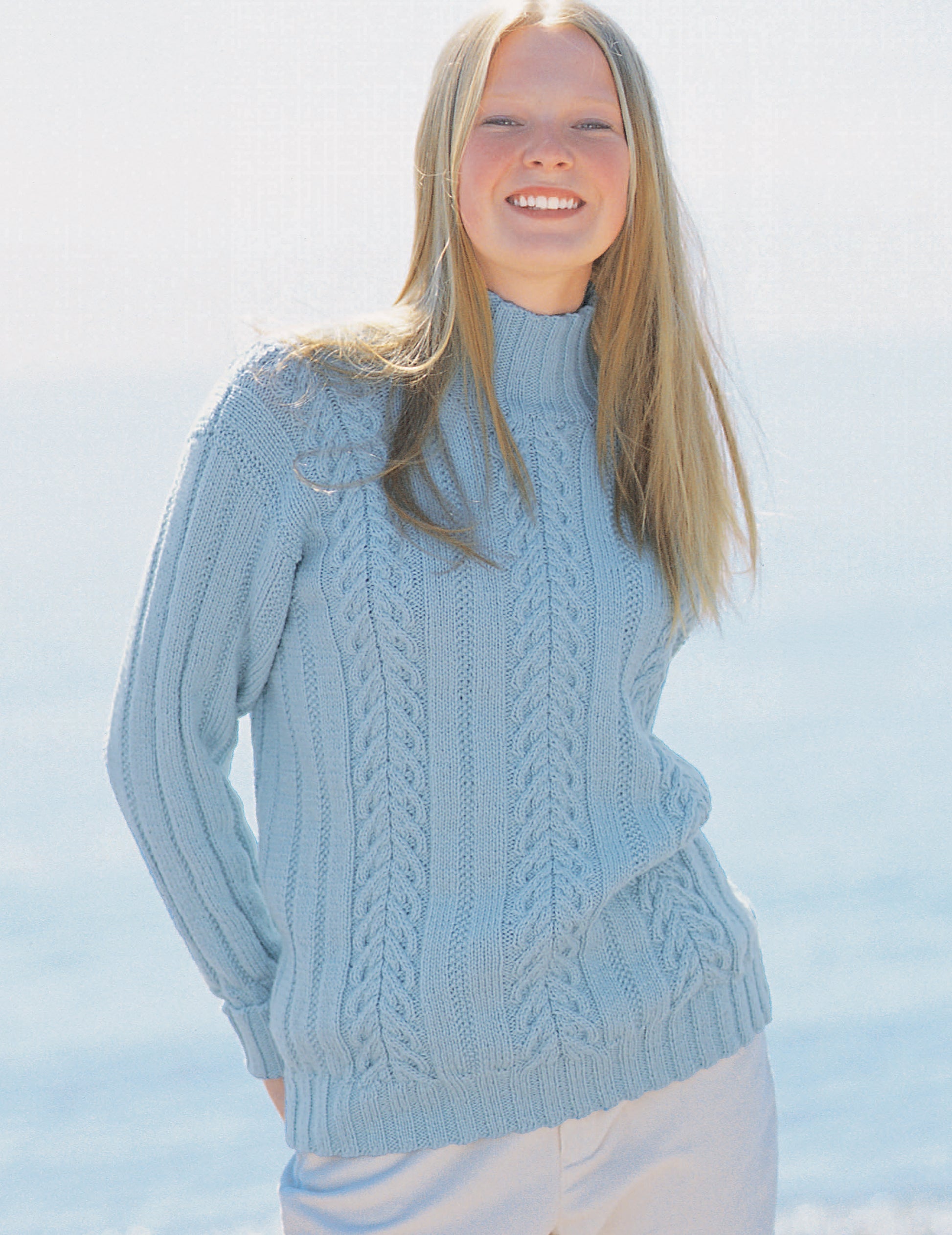 A young woman with long blonde hair smiles outdoors in a light blue cable-knit sweater from The Sweater Collection Knit by Leisure Arts, paired with light pants against a bright, blurred seaside background.
