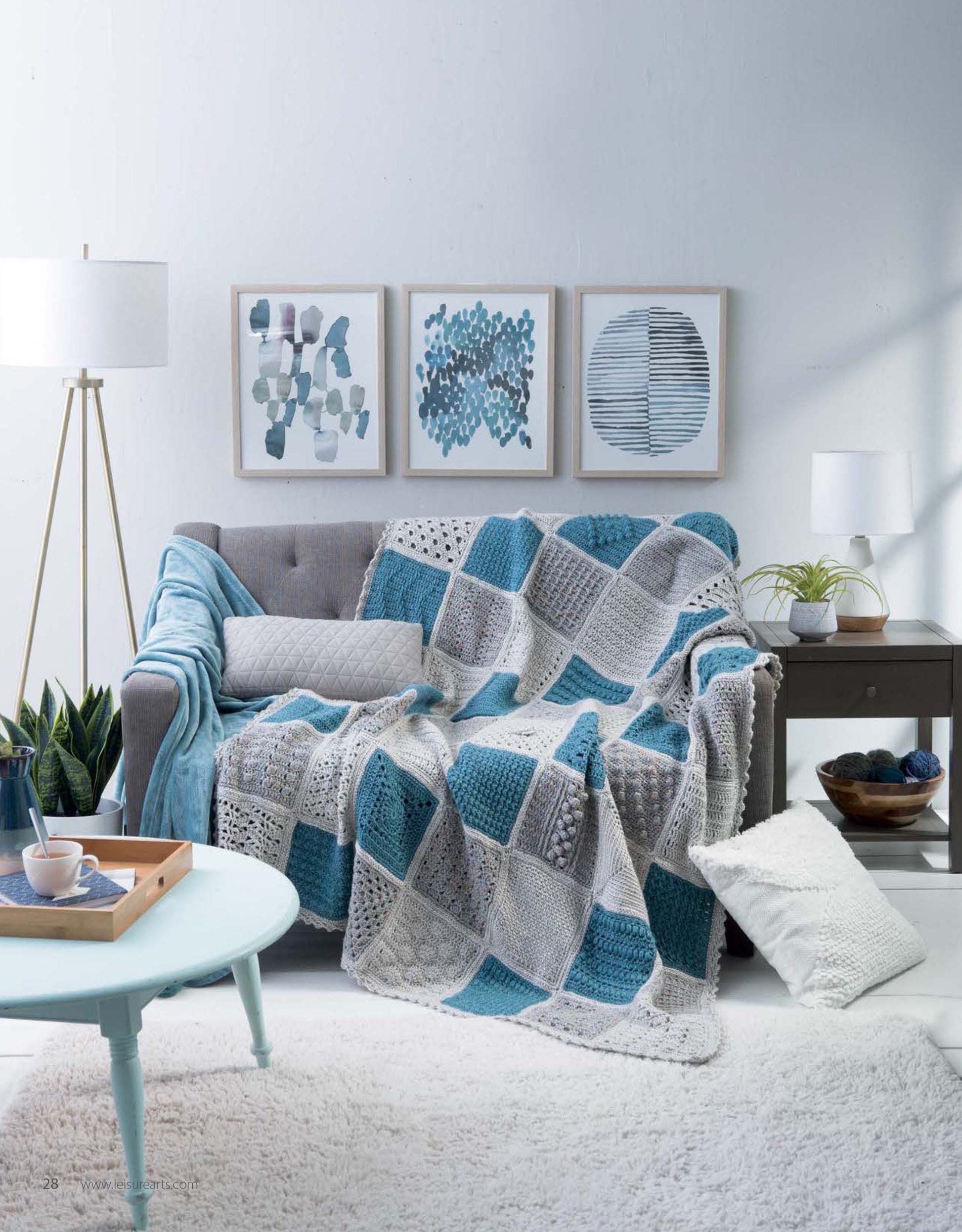 A cozy living room with a gray sofa draped in a blue, white, and gray throw made using Leisure Arts’ Easy to Crochet Pattern Stitches Digital Download. Neutral and blue tones, a white rug, coffee table, potted plants, and abstract art complete the space.