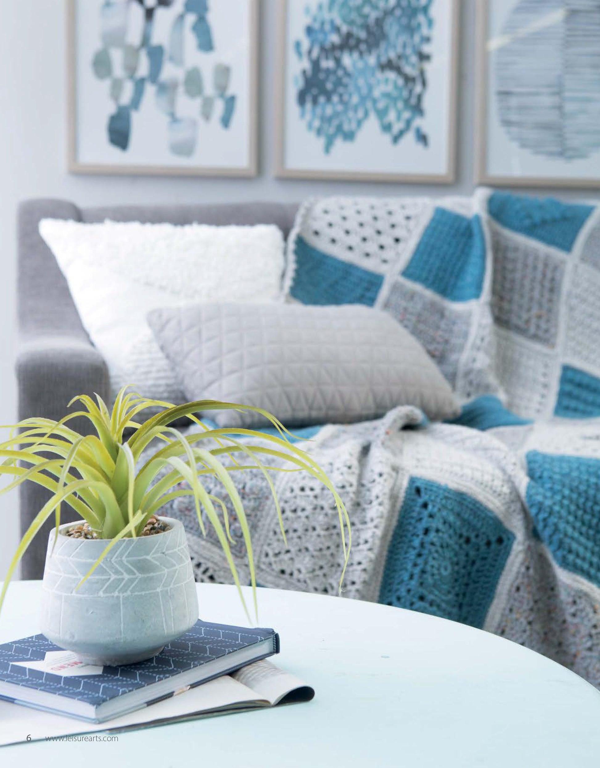 A modern living room features a gray sofa with a blue and gray patchwork blanket made using the Easy to Crochet Pattern Stitches Digital Download by Leisure Arts, plus a gray pillow and white cushion. A potted plant sits on books on a light blue table. Abstract art hangs above.