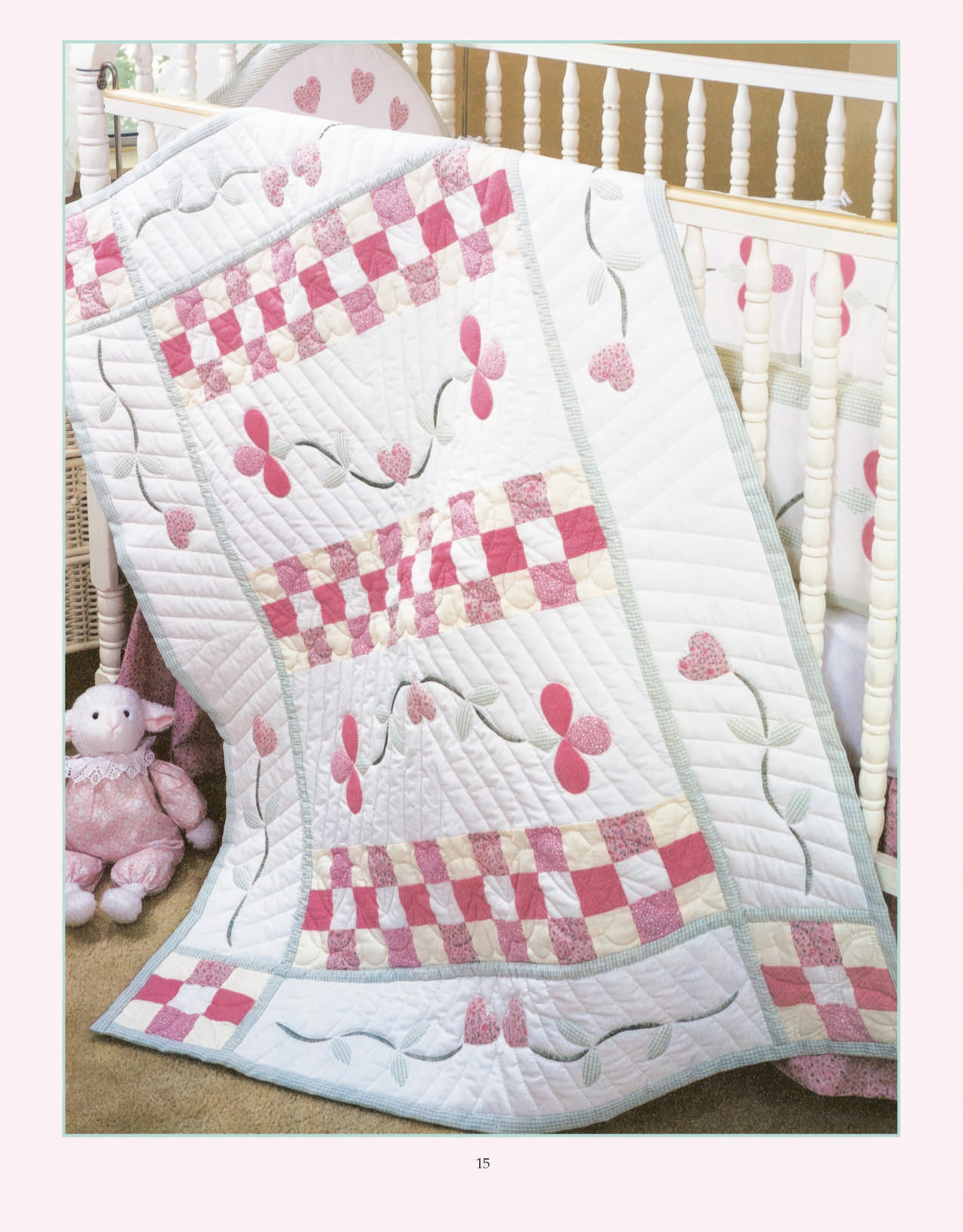 The Rock-a-Bye Quilts Digital Download by Leisure Arts features a pastel pink and white patchwork baby quilt with floral and heart patterns, draped over a crib. A plush pink sheep toy rests on the nearby beige carpet, creating a cozy nursery vibe.