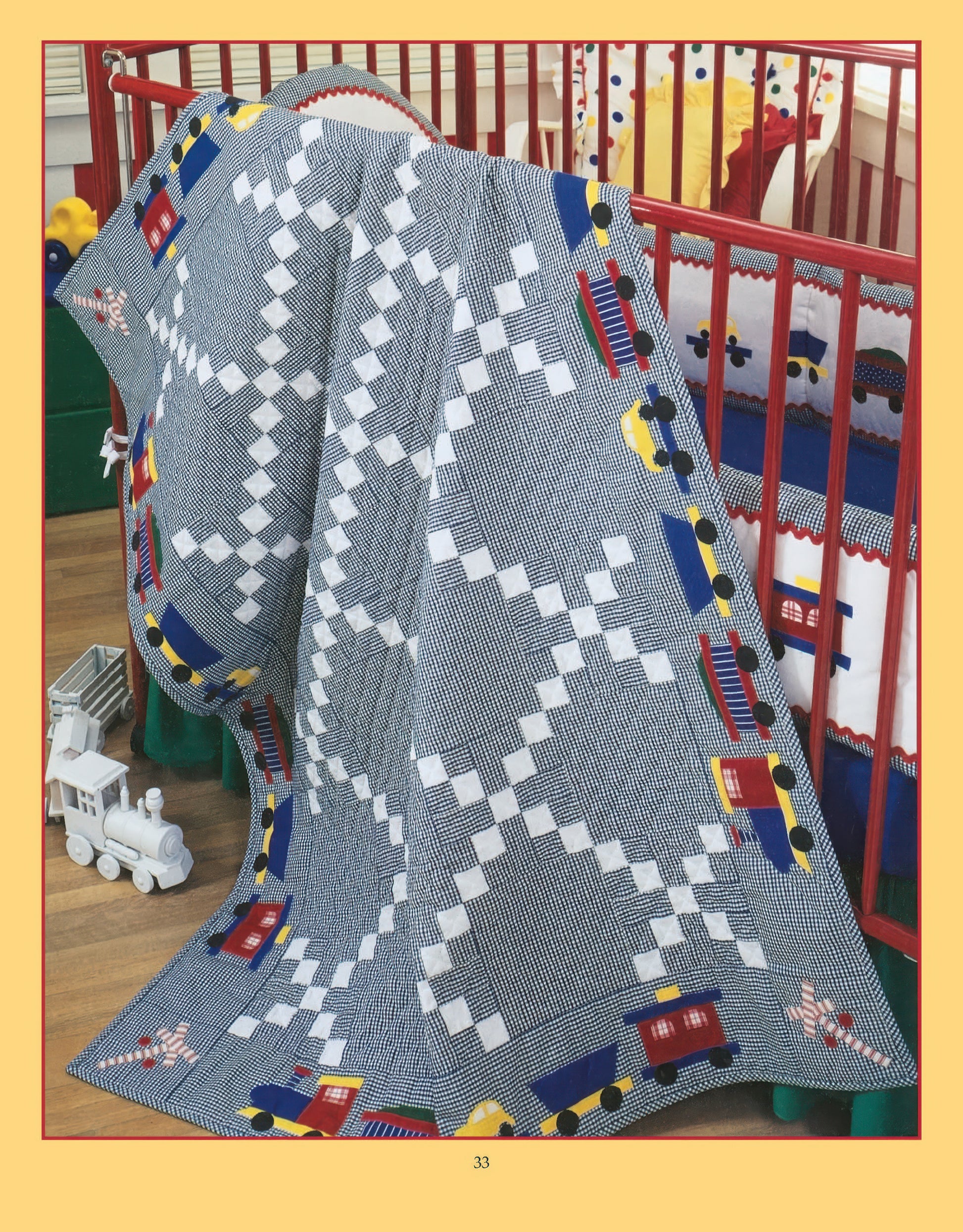 The Rock-a-Bye Quilts Digital Download by Leisure Arts features a blue and white baby quilt with a colorful train motif, displayed on a red crib in the nursery, with a white toy train on the nearby floor.