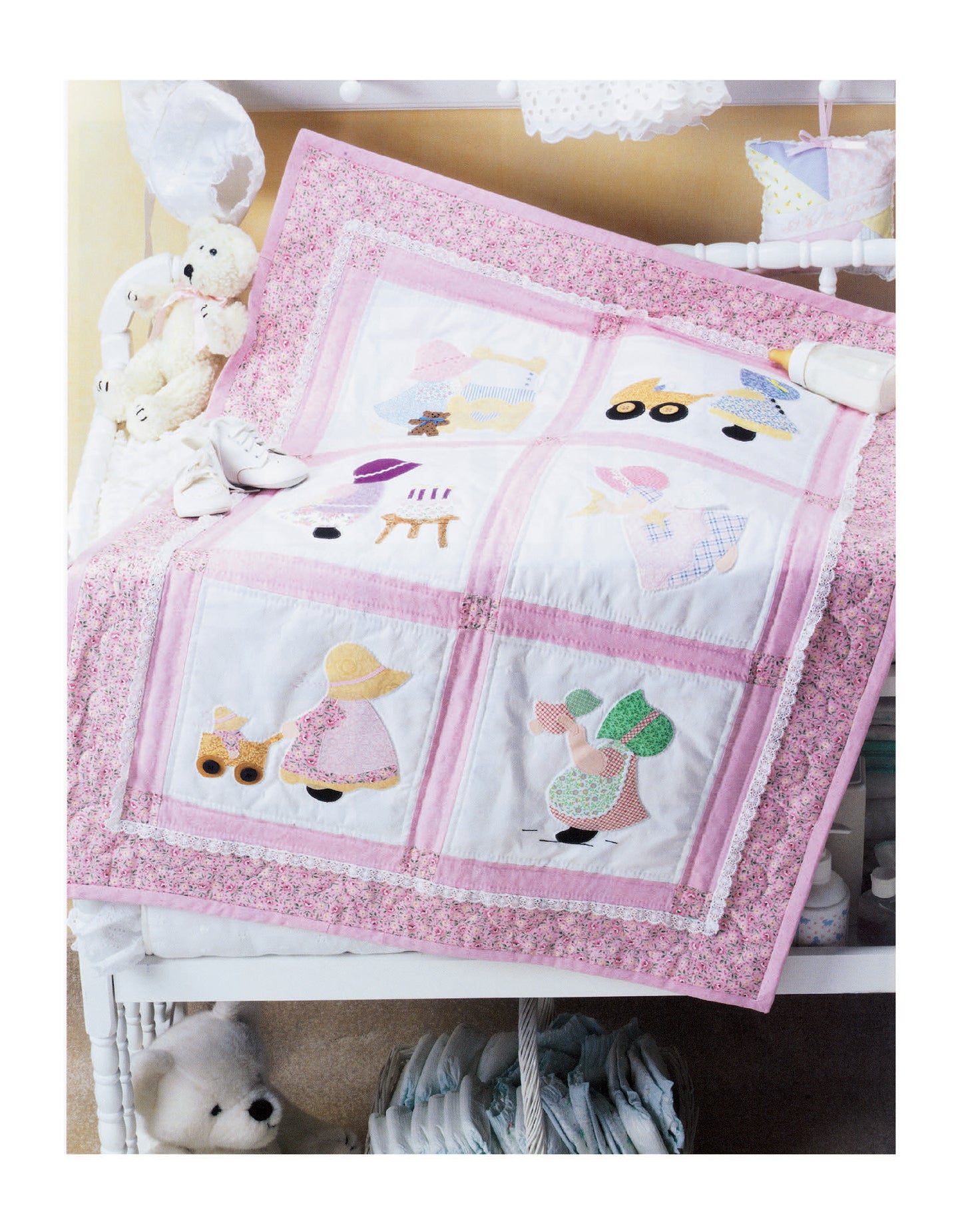 A white crib is draped with a pink and white quilt featuring embroidered children and animals, inspired by classic Sunbonnet Sue applique patterns, made with The Ultimate Sunbonnet Sue Collection Digital Download by Leisure Arts.