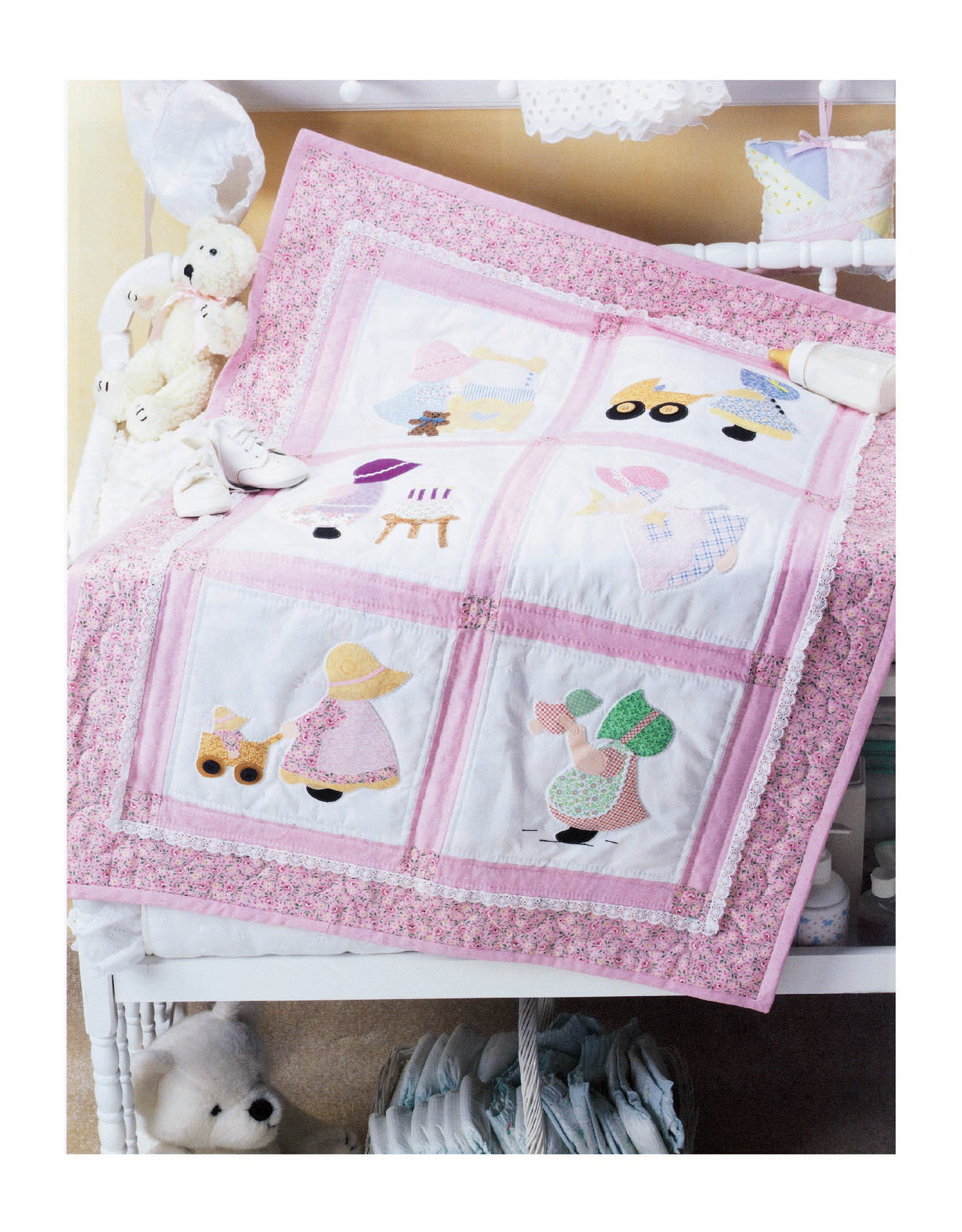A white crib is draped with a pink and white quilt featuring embroidered children and animals, inspired by classic Sunbonnet Sue applique patterns, made with The Ultimate Sunbonnet Sue Collection Digital Download by Leisure Arts.