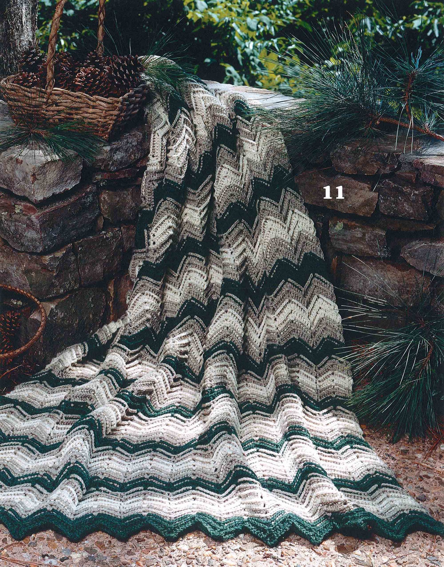 A beginner afghan from Leisure Arts' "Rippling Effects—16 Afghan Designs to Add to Your Home Decor" digital download, made with green, gray, and cream worsted yarn in a ripple pattern, drapes over a stone wall outdoors among pine branches and a wicker basket.